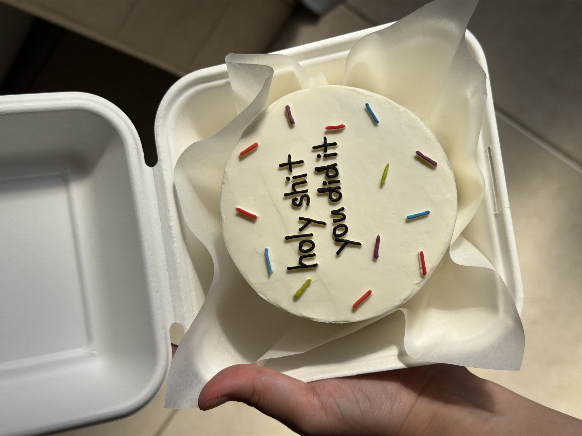 A hand is holding open a white styrofoam takeout container revealing a small, round, white frosted cake. The cake is decorated with multi-colored sprinkles and black frosting that spells out "holy shit you did it". The background is blurred and appears to be an indoor setting. There is no indication this photo was taken in Tel-Aviv, Israel.