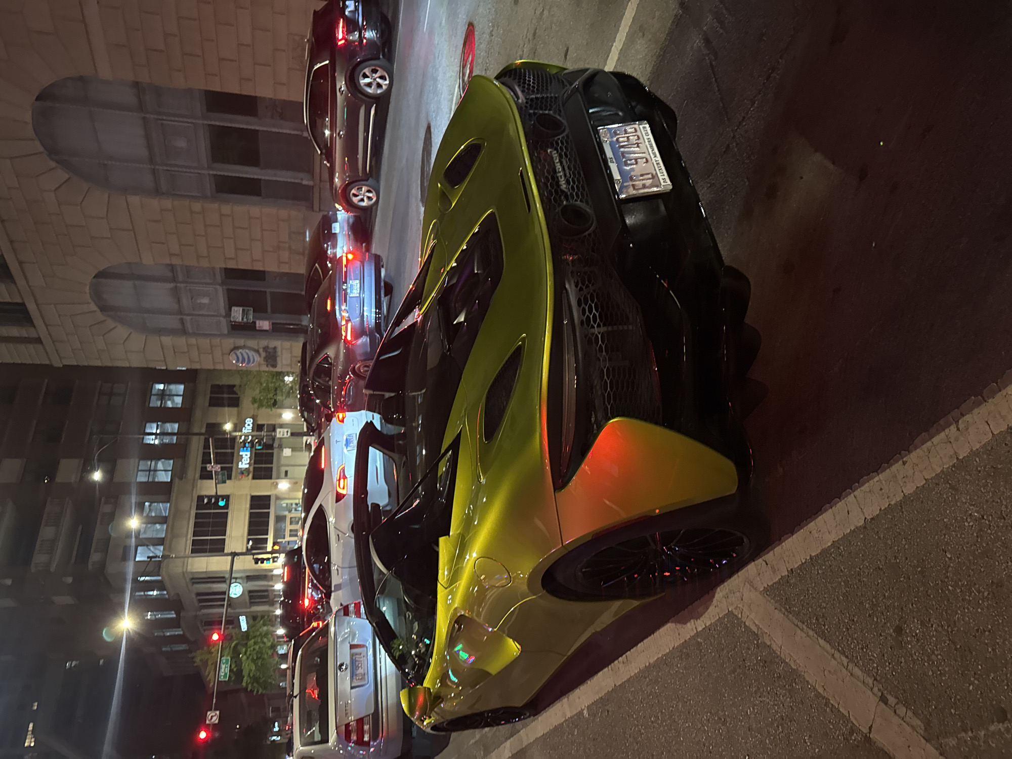 The image depicts a yellow McLaren sports car parked on a street in Chicago at night. The license plate on the car reads "EG 97486." Several other cars are also parked along the street. In the background, there are buildings, streetlights, and a FedEx office sign. The image is taken at street level.