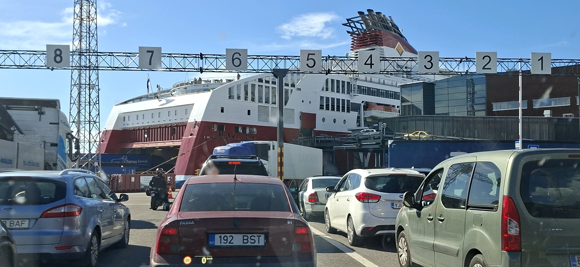 The image depicts a ferry terminal in Helsinki, Finland, under a clear blue sky. Several cars are queuing for boarding, facing a large white and red ferry docked in the background. Numbered signs (8, 7, 6, 5, 4, 3, 2, 1) are visible above the lanes, likely indicating boarding positions. A motorcycle is also present in one of the queues, and a person is seen on it. Various vehicles, including trucks, passenger cars, and a van, are in the scene. The ferry itself is a prominent feature, with its distinctive red hull and white superstructure. The background includes modern buildings with glass facades, suggesting an urban port environment.