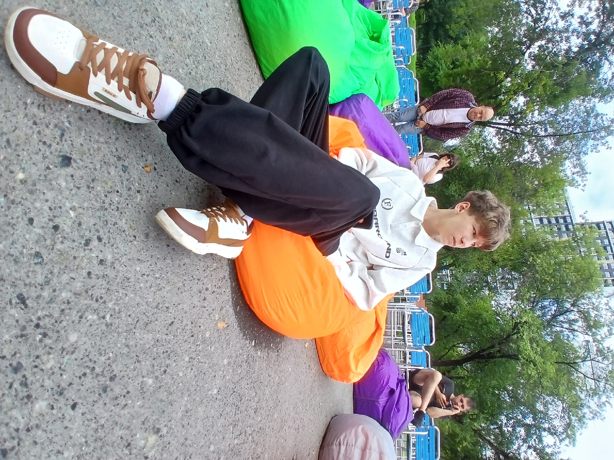 In an outdoor setting with trees and buildings in the background, a young person with light brown hair is seated on an orange beanbag chair, facing to the right. They are wearing a light-colored polo shirt with a logo on the left chest, dark pants, and white and brown sneakers. Other people are visible in the background, including a man in a plaid shirt and jeans standing near some blue chairs, a woman in a white t-shirt and sunglasses, and another man seated on a purple beanbag chair, looking at his phone. Several large beanbag chairs in green, purple, and orange are arranged on a paved ground. The overall scene suggests an outdoor event or gathering in a park-like environment. The presence of tall apartment buildings in the distance and the style of the blue chairs suggest a location within the city of Yekaterinburg, Russia.