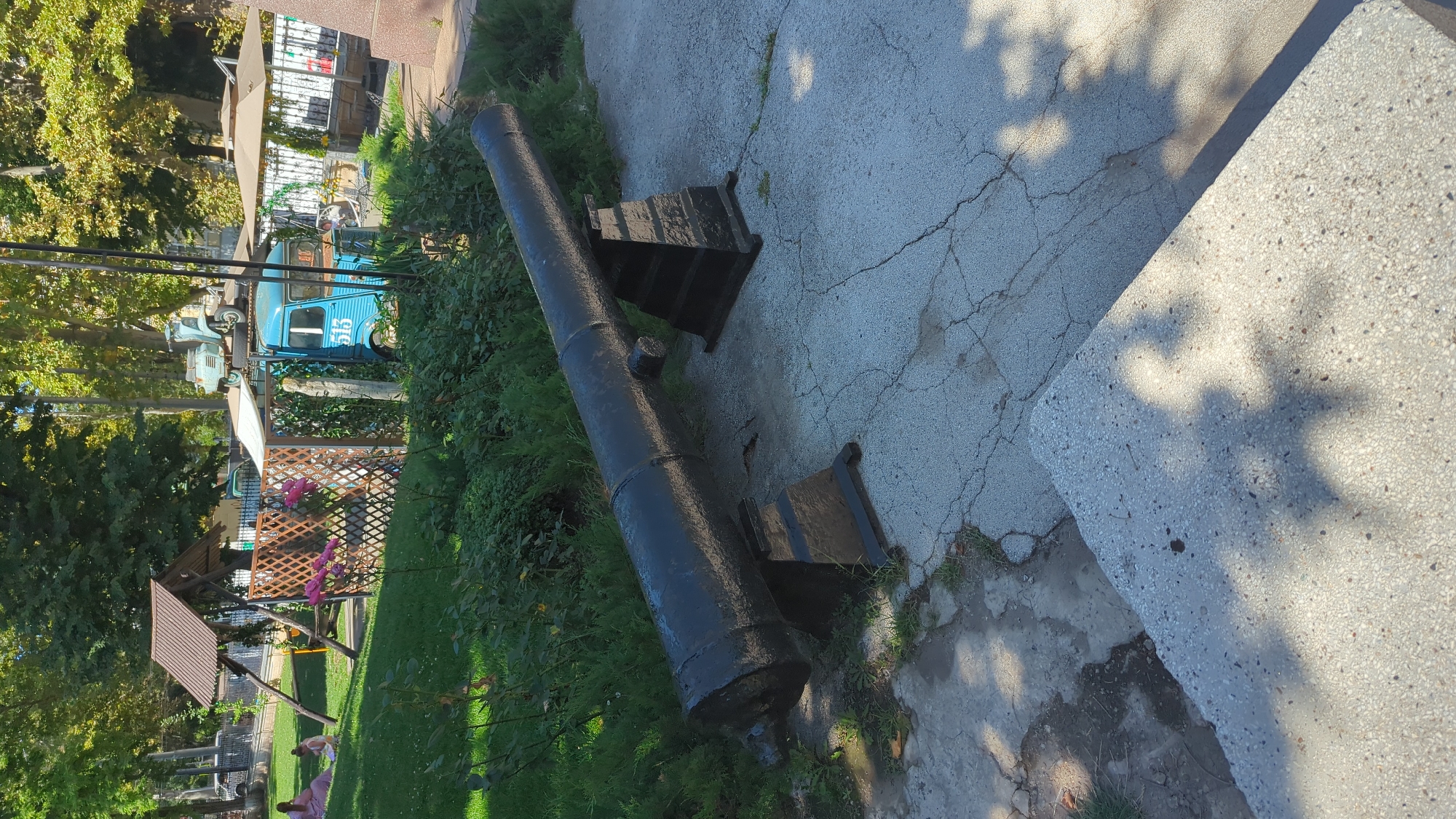 A weathered black cannon rests on a concrete platform in what appears to be a park or public space. The cannon is positioned on two black metal stands. The background reveals a green grassy area with trees, a small wooden playground structure, and a blue retro van with the number "513" prominently displayed. The scene is bathed in sunlight, casting shadows on the ground. Two individuals, possibly women, are visible in the distance sitting on the grass. The environment suggests a recreational area, possibly in Chișinău, Moldova, given the presence of the distinctive blue van.