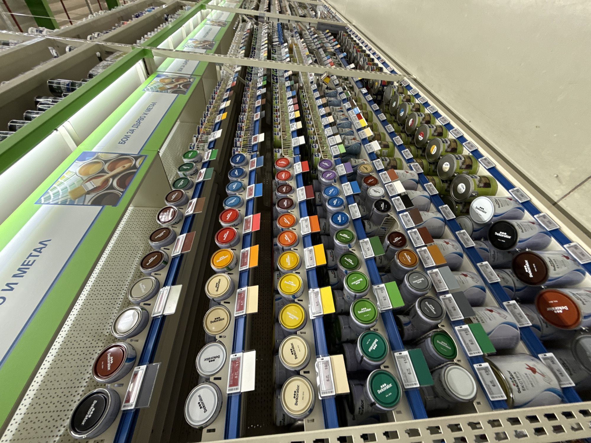 The image shows shelves stocked with rows of paint cans of various colors inside a retail store. The cans, labeled "Dekorator" in Cyrillic, are arranged vertically on blue shelving, with color swatches affixed to the front of each can. Signage in Bulgarian reads "Бои за дърво и метал" ("Paints for wood and metal"). The perspective is angled upwards, creating a sense of depth and height, suggesting a wide selection of paints is available. The location is likely a hardware or home improvement store in Stara Zagora, Bulgaria.