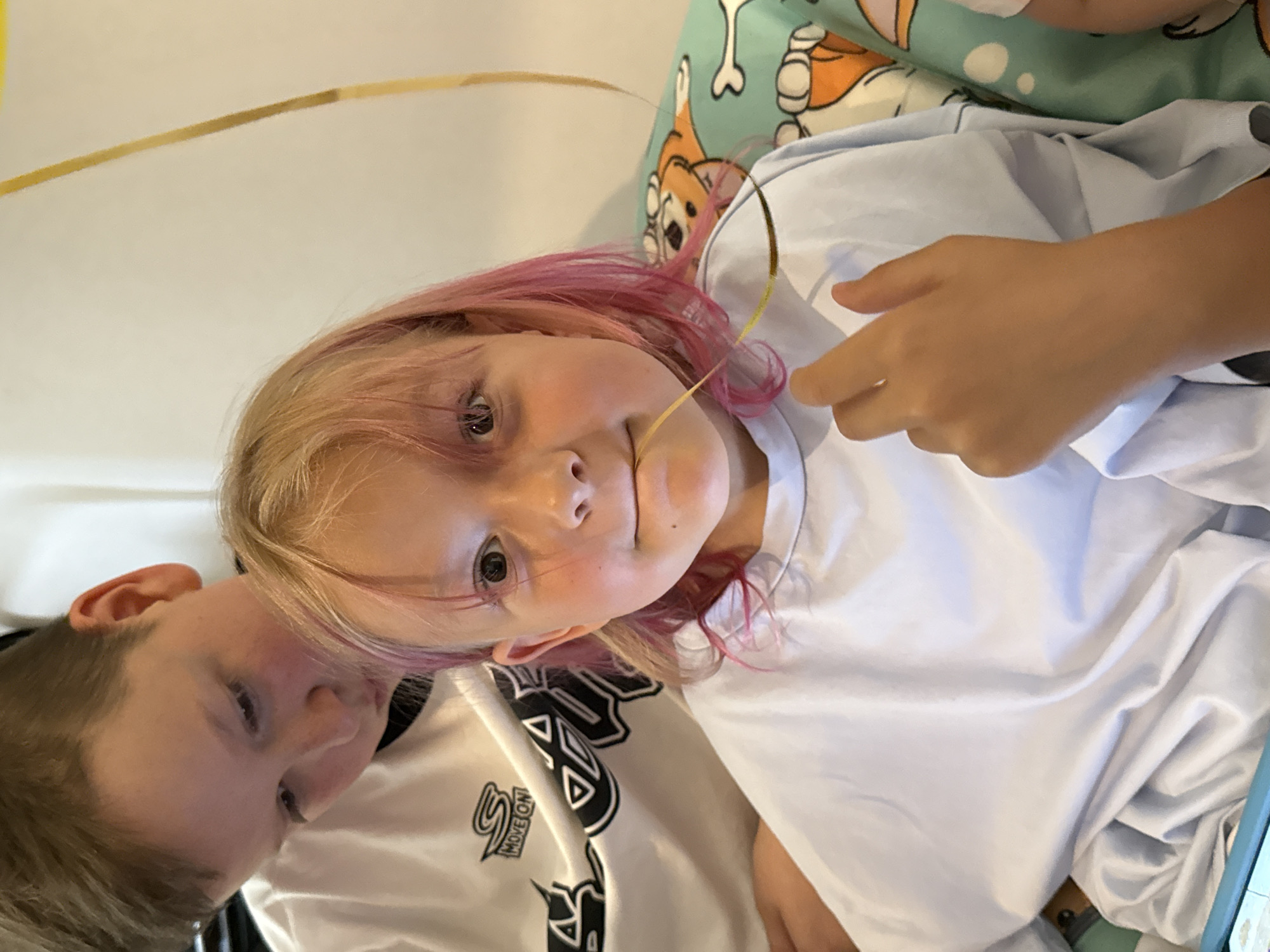A young girl with blonde and pink hair is featured prominently in a close-up, slightly tilted portrait. She has light skin and is looking directly at the viewer with wide, dark eyes. Her mouth is slightly open, revealing a hint of a smile or a playful expression. A thin, golden-yellow strand, possibly a party streamer or a piece of decor, is draped across her face, with one end near her mouth and the other extending upwards and to the right, held lightly by her right hand.
She is wearing a white t-shirt with visible folds. To her left, partially visible, is another individual, likely a boy, wearing a white t-shirt with black graphics that appear to say "SMOKE ON" or "MOVE ON." Only the side of his face and ear are visible, and he seems to be looking down or away from the camera.
In the background, behind the girl's head, there's a pillow with a light blue or teal cover featuring cartoon-like dog characters, one of which appears to be a corgi. Another pillow or blanket with a lighter green color and white polka dots is also visible. The lighting is bright and even, suggesting an indoor setting. The overall mood appears casual and playful. There are no clear indications of the time of day or specific location.