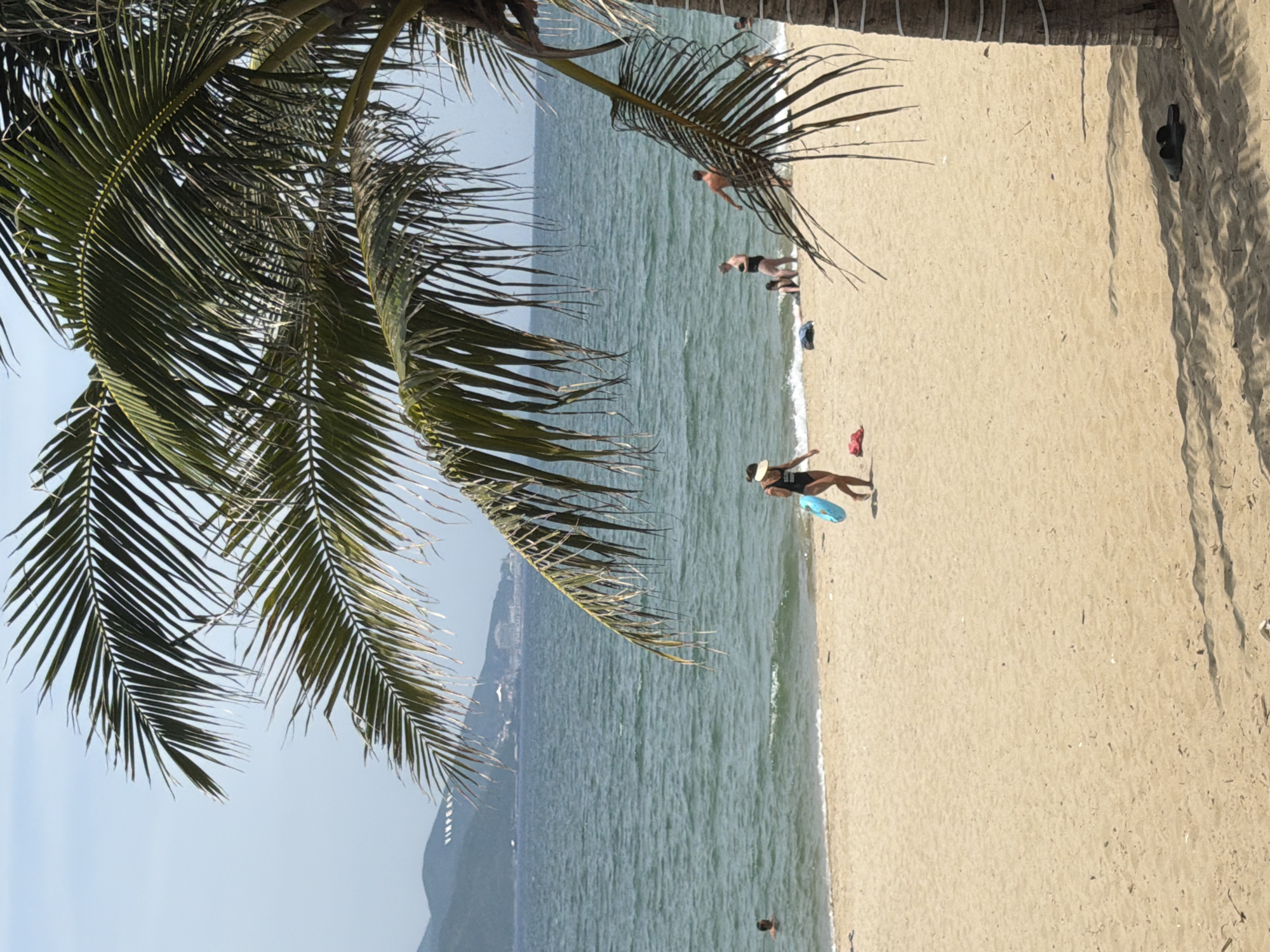 A woman in a black swimsuit and a white visor walks along a sandy beach in Nha Trang Ward, Vietnam, carrying a blue inflatable ring. The ocean is to her left, with a man swimming in it. Further to the right, a man and child are in the shallow water, and another man is standing on the beach. The scene is framed by the fronds of a palm tree. In the distance, hills with buildings are visible. The sand is lightly shadowed, indicating it is daytime.