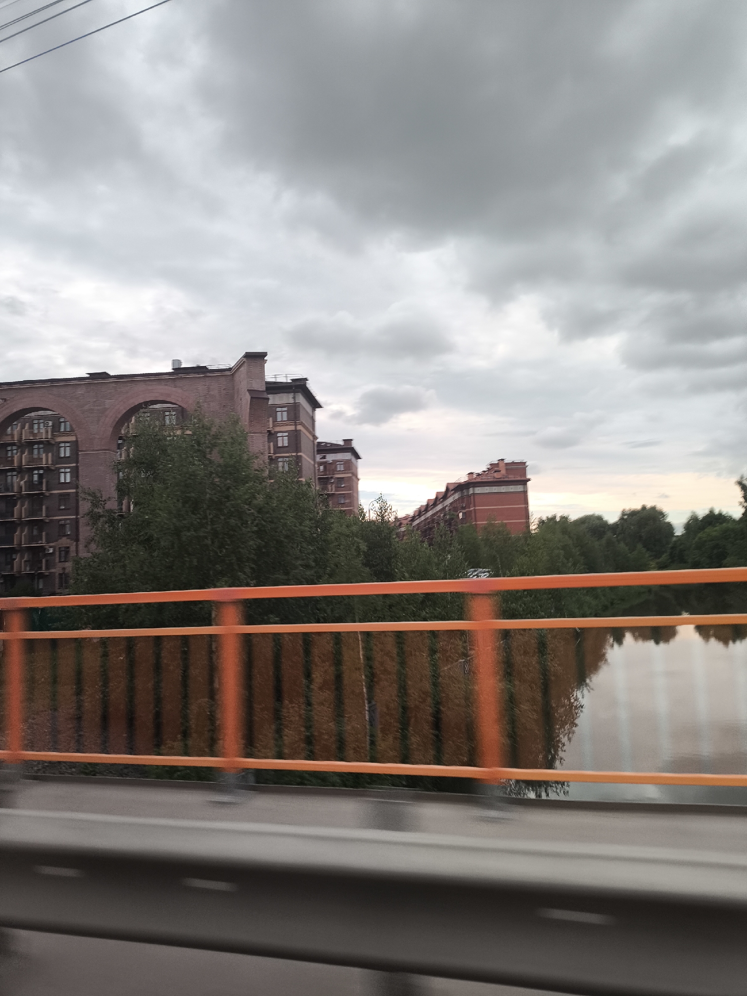 A side view shows an overcast sky above a river and a row of brick apartment buildings. In the foreground, an orange railing on a bridge is visible. The reflection of the railing and trees can be seen in the water. The apartment buildings are tall and modern, with many windows. Trees are scattered throughout the scene, some in front of the buildings and some along the riverbank. The overall mood of the image is calm and slightly muted due to the cloudy weather.
