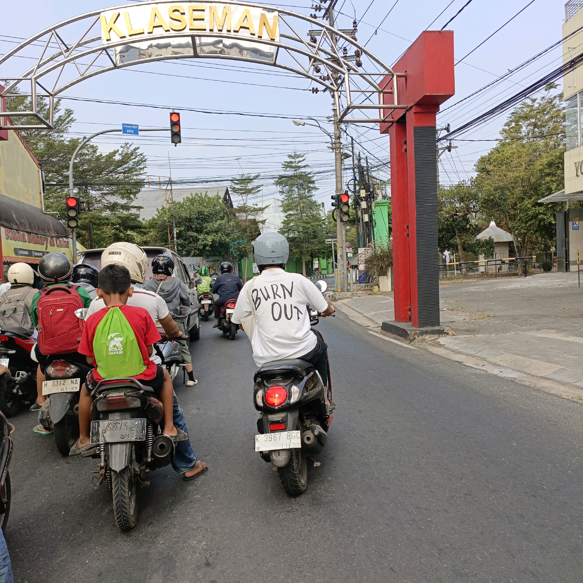 The image captures a street scene in Salatiga, Indonesia, likely in the vicinity of the "KLASEMAN" archway. The focus is on several motorcyclists in traffic, with a prominent red and black pole structure on the right side of the frame. Traffic lights are visible and illuminated red, indicating a stop.
In the foreground, a young person in a red shirt and a green backpack with an elephant logo is riding on the back of a motorcycle. The driver of this motorcycle, wearing a helmet, is partially visible. To their left, another motorcyclist with a red backpack and helmet is also stopped. Further ahead, other motorcyclists and a car are visible, also stopped at the red light.
The person in the immediate center, riding a black scooter, wears a white t-shirt with the words "BURN OUT" printed on the back in black letters. They are also wearing a grey helmet.
The environment includes buildings, trees, and a complex network of electrical wires overhead. The road surface appears to be asphalt. The overall impression is a typical urban traffic situation in Indonesia.