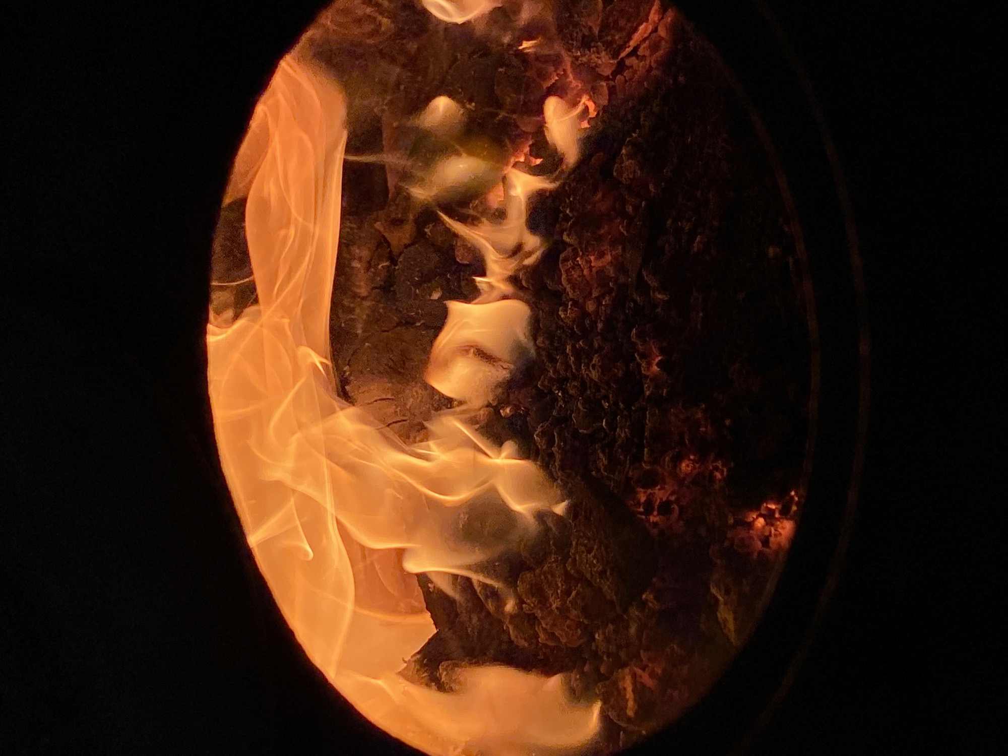 The image displays a vibrant, active fire viewed through a distinct circular opening. Bright, flowing flames dominate the left and center of the circular frame, exhibiting warm hues of orange, yellow, and red. These ethereal flames appear to be consuming or dancing around a rough, textured mass of burning material, which resembles charred wood or embers, visible in the center and right side of the circle. Portions of the burning material glow with intense red and orange light. The area immediately outside the circular opening is uniformly dark black, suggesting an enclosed space like a wood stove, a fireplace, or a fire pit at night, where the fire is the sole source of illumination. The edges of the circular opening itself are dark and form a clear boundary between the fire and the surrounding darkness. No people or visible text are present in the image. The scene evokes a sense of warmth and coziness.