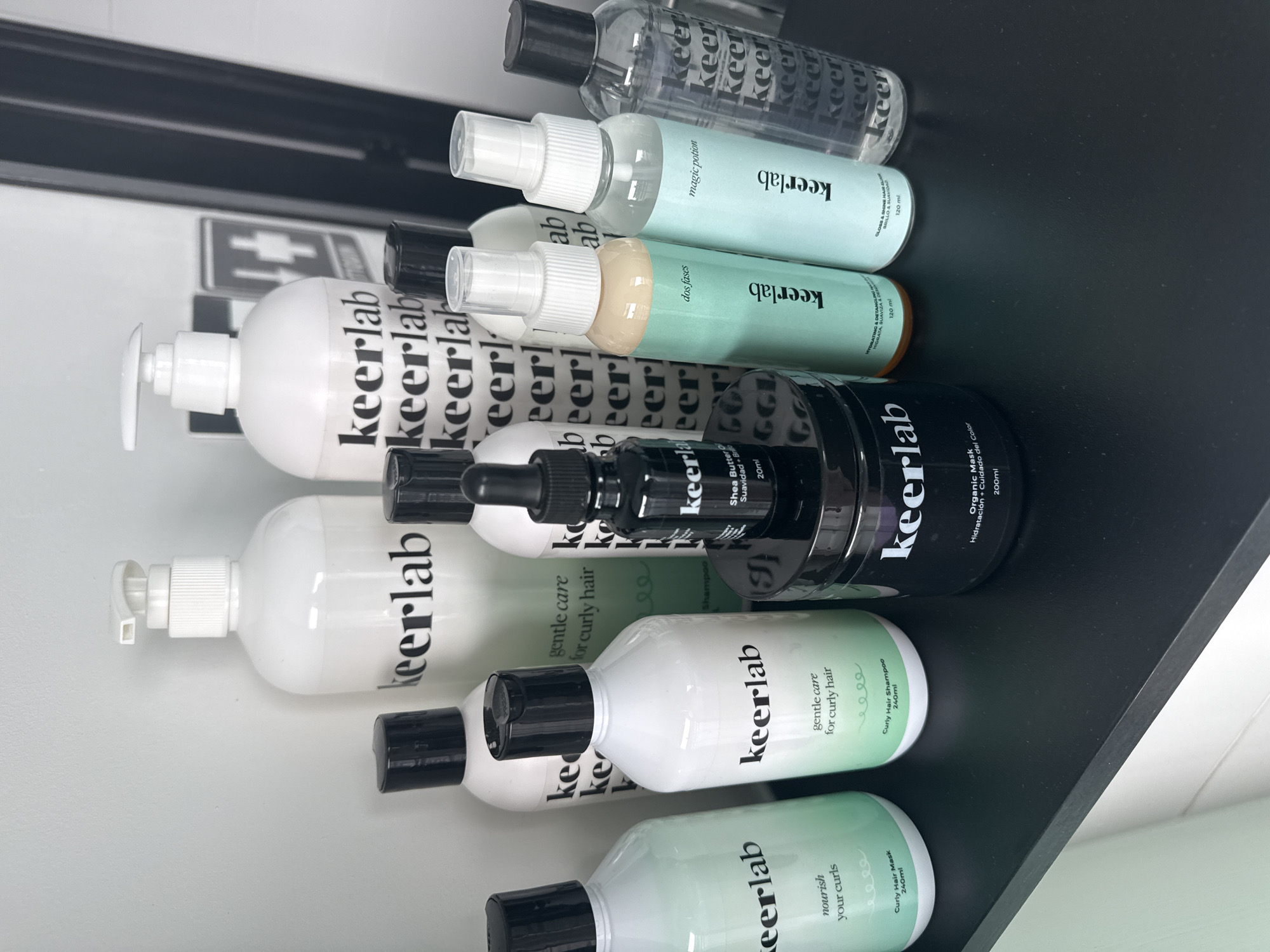 A collection of hair care products from the brand "keerlab" is neatly arranged on a dark, reflective surface, likely a shelf or countertop, in what appears to be an indoor setting. The image is taken from a slightly elevated, oblique angle, looking down at the products.
The array includes various types of bottles and containers: several white pump bottles, clear and light-green spray bottles, a small black dropper bottle, and a larger black jar. The majority of the packaging is white with black lettering, while some bottles feature a light green or teal color, suggesting different product lines or ingredients.
Prominently displayed on all items is the brand name "keerlab." Visible product descriptions include: "magic potion," "dos fases" (two phases), "GLOSS & SHINE HAIR BLEND," "HYDRATING & DETANGLING," "gentle care for curly hair," "nourish your curls," "Curly Hair Shampoo 240ml," "Curly Hair Mask 240ml," "Shea Butter 20ml," and "Organic Mask Hidratación + Cuidado del Color 200ml." Some bottles also feature a repeating pattern of the "keer" logo.
In the blurred background, behind the products, a light-colored wall or surface is visible, along with a faint grey symbol that resembles a first aid cross. No specific activity is occurring; the scene depicts a static display of hair care products. The lighting is even, suggesting an indoor environment without strong external light sources.