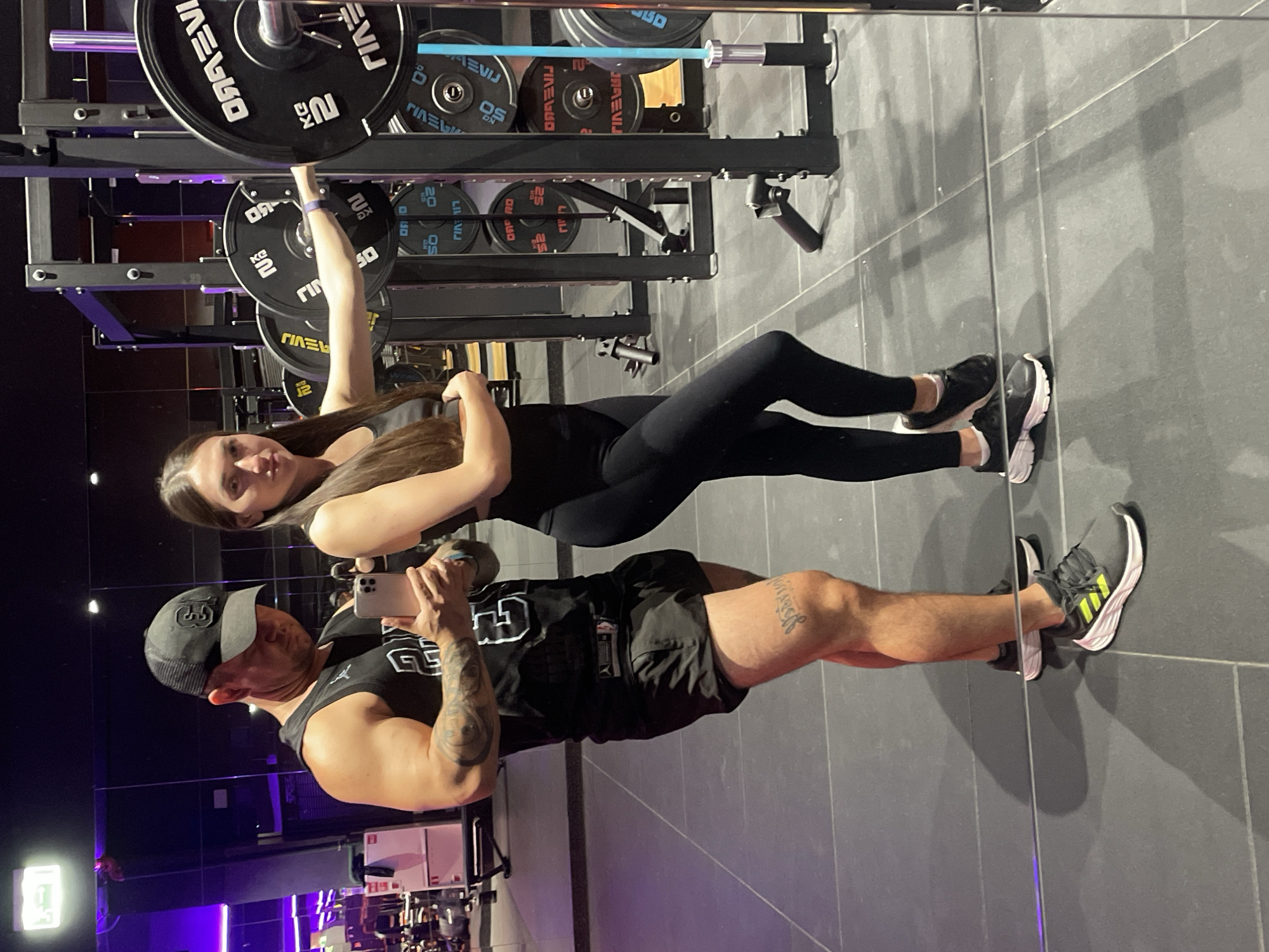 The image depicts a couple in a gym. The man, wearing a black baseball cap and a black tank top, is holding a smartphone and looking at it. He has tattoos on his arm and leg. The woman, wearing black leggings and a black sports bra, is standing next to him, her hand resting on a weight rack with large circular weights. The gym has a dark, tiled floor and mirrors, with gym equipment visible in the background. The lighting is dim with some purple accents. There is no indication that this location is in Kazan, Russia.