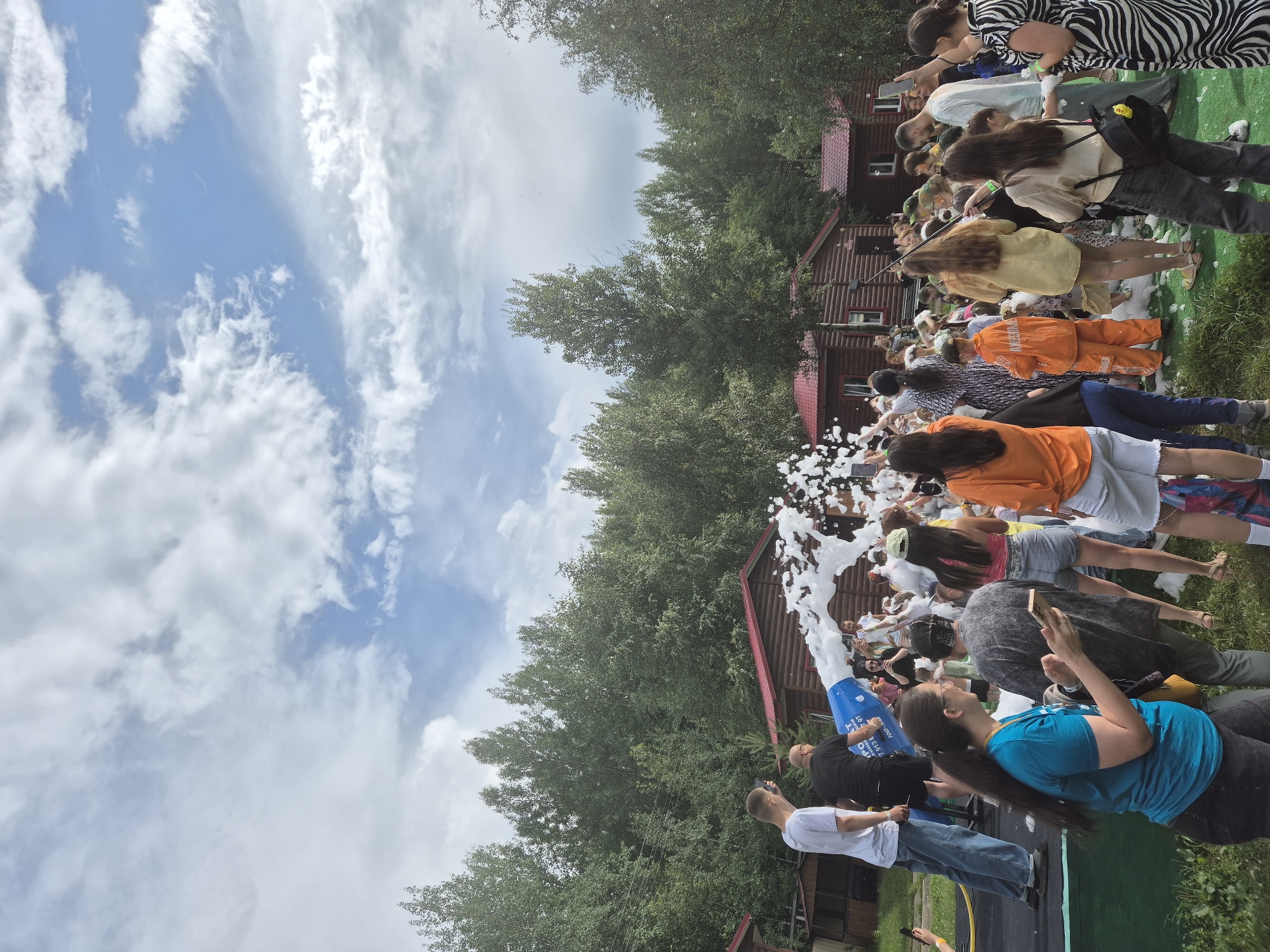 In Nizhnevartovsk, Russia, a foam party is underway. A large foam machine is spraying a torrent of white foam into the air, creating a frothy spectacle. A crowd of people, dressed in casual summer attire, are gathered around the machine, some actively participating in the foam party and others observing. The scene is set outdoors, with a backdrop of lush green trees and rustic wooden cabins, suggesting a park or recreational area. The sky above is partly cloudy, with patches of blue peeking through. The overall atmosphere appears festive and joyful, with individuals of various ages enjoying the unique experience.