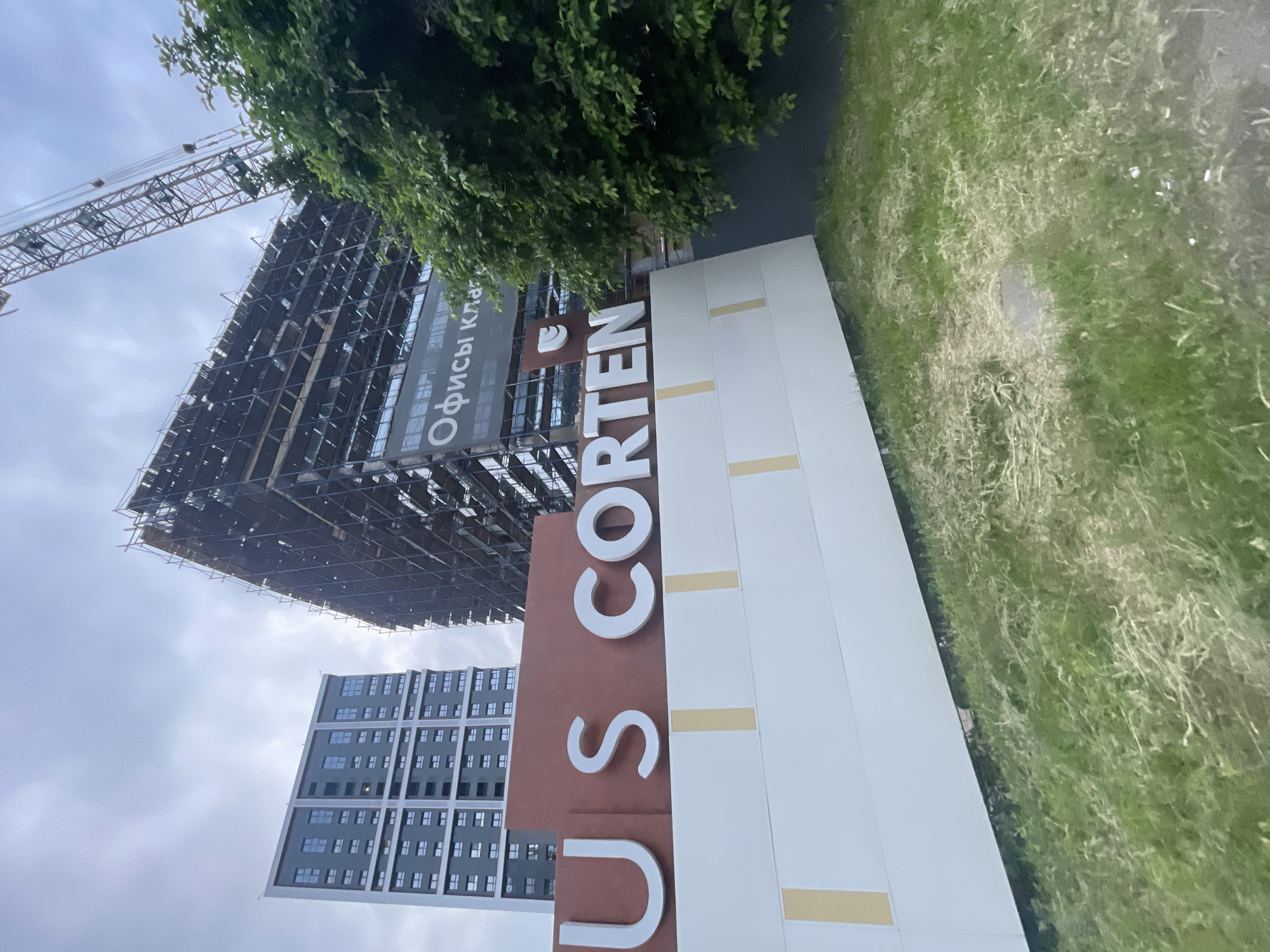 The image displays an urban landscape under an overcast sky, featuring a large, multi-part sign in the foreground and two tall buildings in the background. The foreground sign has a reddish-brown upper section with prominent white, capitalized letters spelling "U S CORTEN." Below this is a white segmented base with vertical light beige strips. The ground beneath the sign is an unkempt grassy area with patches of dried vegetation.
To the left, a tall, modern building with a dark gray facade and numerous rectangular windows appears completed or near completion. To the right, a large building is under active construction, extensively covered in dark scaffolding. A tall construction crane extends diagonally upwards and to the right from this building. A banner is visible on the construction site, partially obscured, displaying the Cyrillic text "Офисы класса" (Ofisy klassa), which translates to "Class A Offices." A dense green tree with small leaves partially obstructs the view of the construction building on the far right. No people are visible in the scene. The location is in the city of Unknown, Unknown.