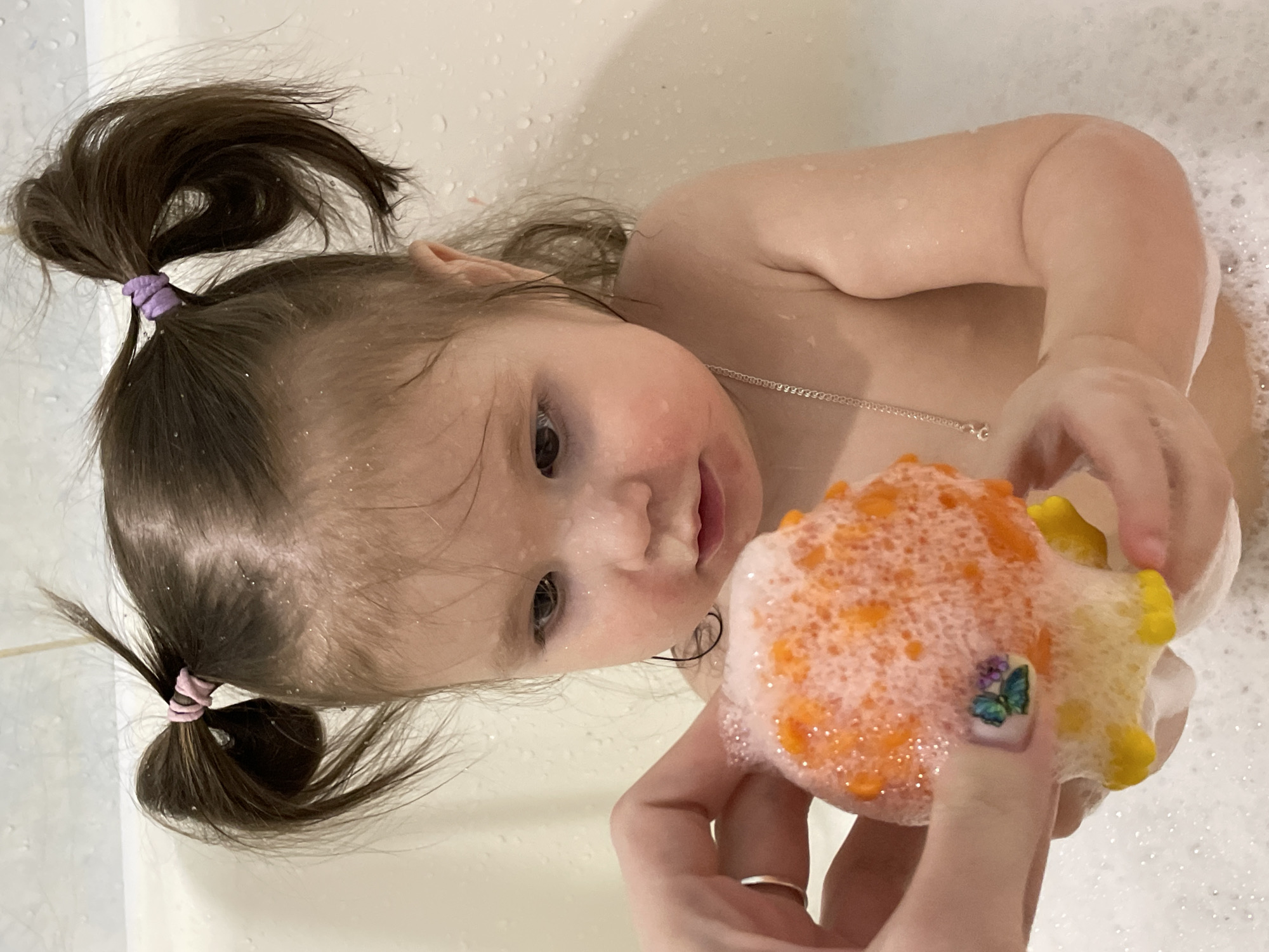 The image depicts a young child, approximately 1-3 years old, bathing in a white bathtub in Izhevsk, Russia. The child has wet, dark brown hair styled in two pigtails, secured with a pink elastic band on the viewer's left and a purple elastic band on the right. Water droplets are visible on the child's hair, face, and the white tiled wall in the background. The child's cheeks appear flushed. A thin silver chain is worn around the child's neck. The child is holding a buoyant, orange and yellow bath toy, which is extensively covered in white soap suds. An adult hand, featuring a silver ring on the fourth finger and nail art depicting a butterfly and floral elements, is also present, engaged in a joint interaction with the child over the toy. The bathtub contains a significant volume of white foam.