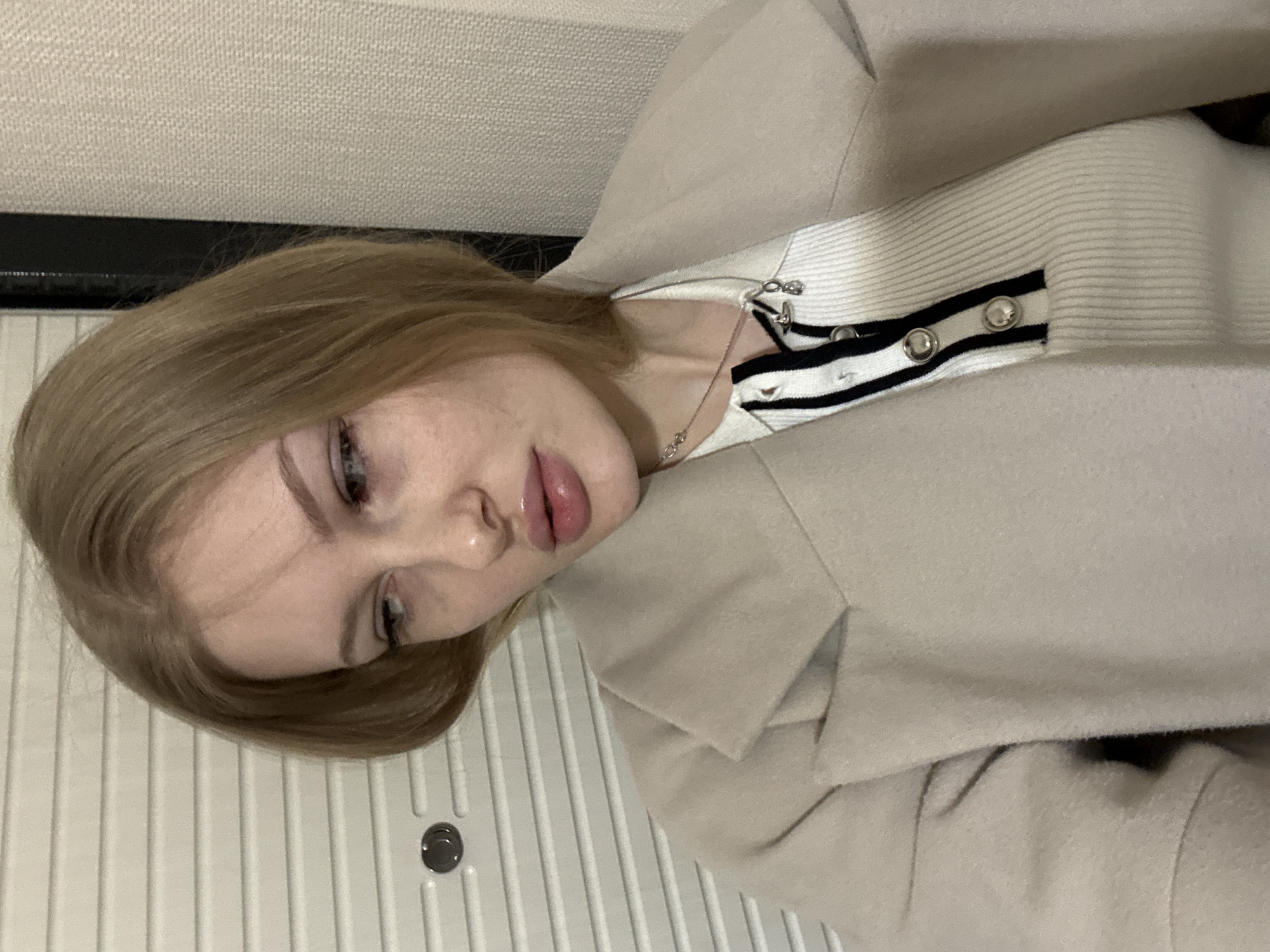 A young woman is posing for a selfie in an indoor setting. She has fair skin, blonde hair, and is wearing light-colored clothing, including a jacket or coat. She also wears a necklace. The background consists of a wall, and a surface with vertical ridges and a round detail that may be a handle or knob.
The location is in Perm, Russia.