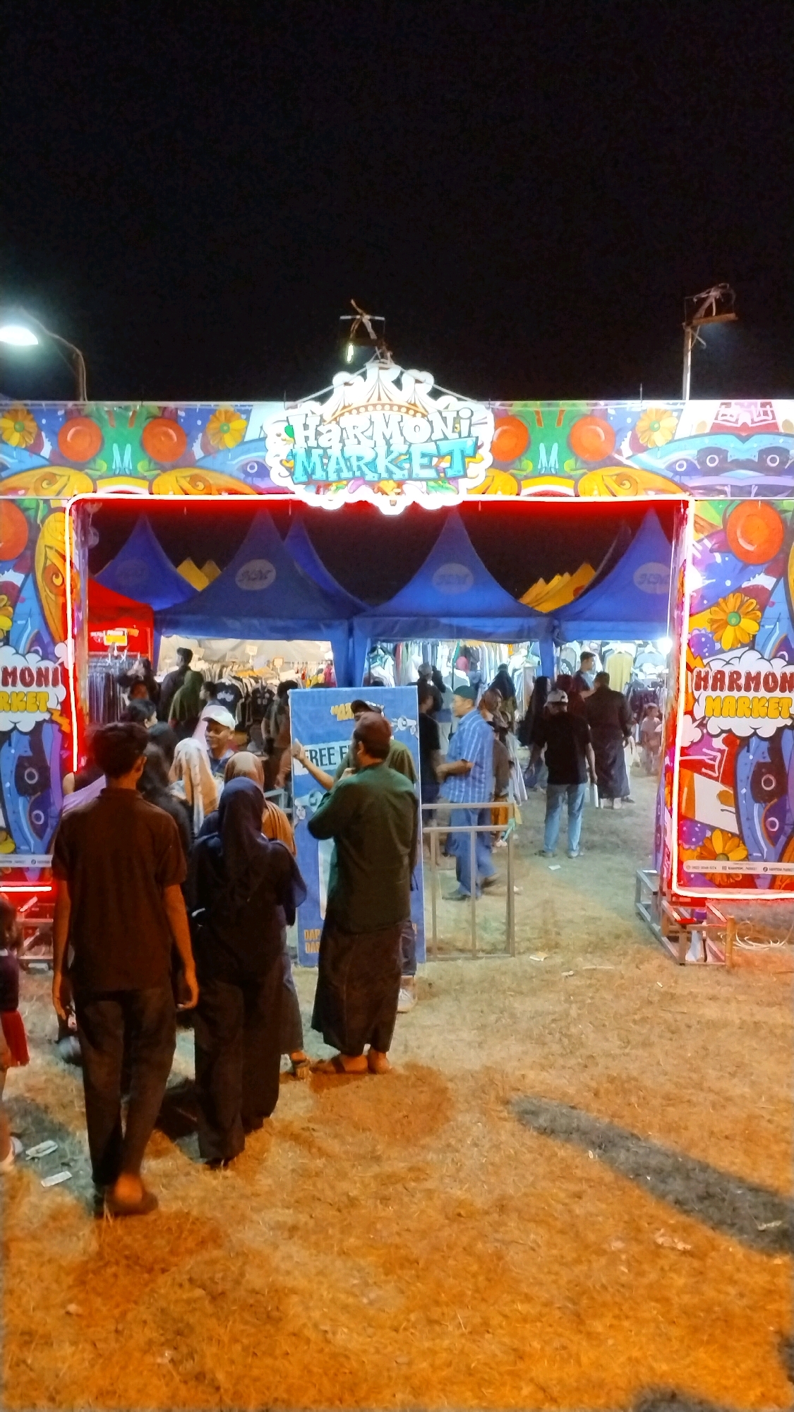 A night scene shows an entrance archway to a market in Canggu, Indonesia. The archway is decorated with colorful murals of fruits and flowers and reads "HARMONI MARKET" in large, glowing letters. Underneath the archway, a blue tent structure houses stalls displaying merchandise, with several people milling about. In the foreground, a group of individuals stands on a dirt-covered ground, some walking and others observing the market entrance. The overall atmosphere suggests a vibrant evening market or festival.