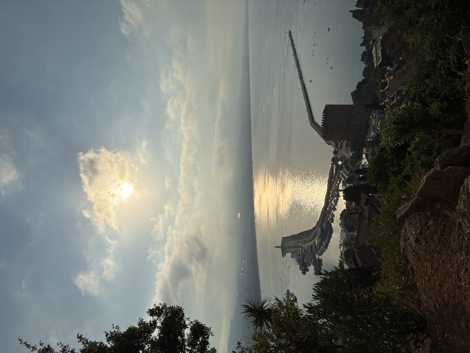 The image is a landscape photograph taken from an elevated viewpoint overlooking the city of Alanya, Turkey, during a sunset. The sun is low in the sky, partially obscured by clouds, casting a golden, shimmering reflection on the calm sea.
In the foreground, lush green trees and foliage frame the scene, with a rough, rocky terrain visible at the bottom. The middle ground reveals a section of the city with terracotta-tiled roofs nestled amongst the greenery. A prominent red-brick historical structure, likely a castle or fortress, stands on a rocky outcrop overlooking the water.
The foreground extends to a curved pier extending into the sea. At the end of this pier is a lighthouse, and alongside it, a harbor filled with numerous docked boats. Another, longer pier stretches further into the water, lined with streetlights. In the distance, the coastline of Alanya stretches out, with buildings and hills fading into a hazy, atmospheric backdrop. Several small boats are visible on the water, and a few people can be seen on the longer pier. The sky above is a blend of pale blue and soft, white clouds, with the setting sun creating a bright glow. The overall impression is one of serene coastal beauty at dusk.