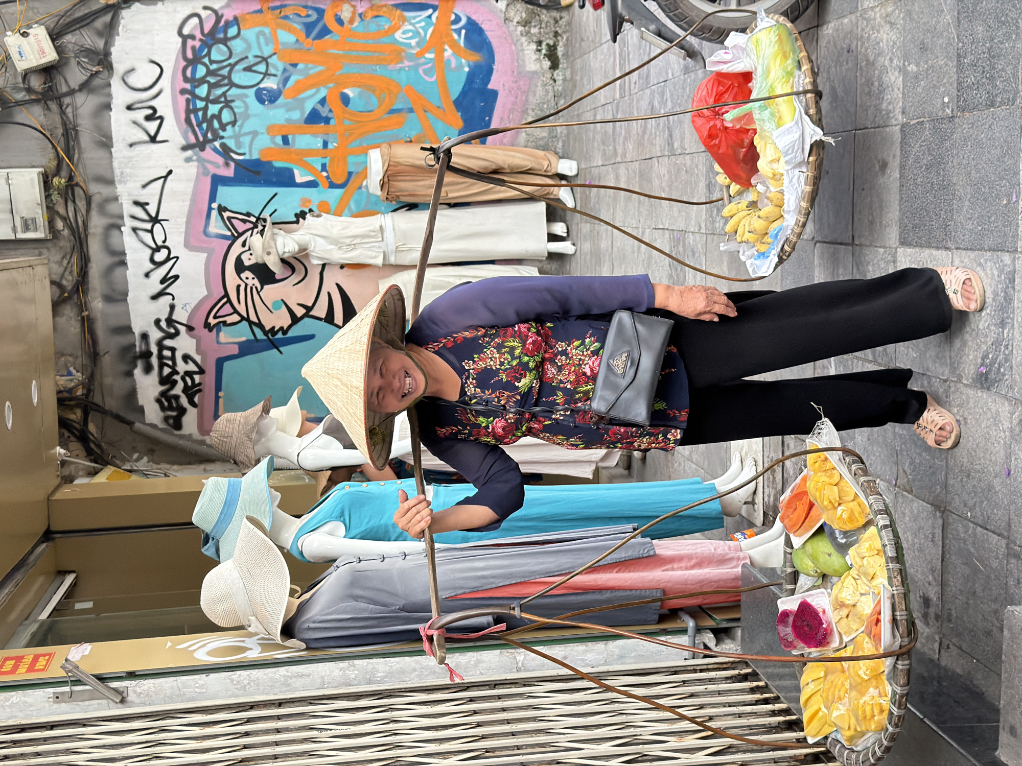 A smiling woman wearing a conical hat is centered in the image. She carries a shoulder pole with baskets of fruits and goods on either end, including bananas, mangoes, and jackfruit. She has a gray bag slung over her shoulder. Behind her, the front of a shop displays mannequins wearing clothes and hats. Graffiti is visible on the wall above the shop. The location appears to be a street in Hà Nội, Vietnam, based on the traditional conical hat worn by the woman and the style of the street scene.