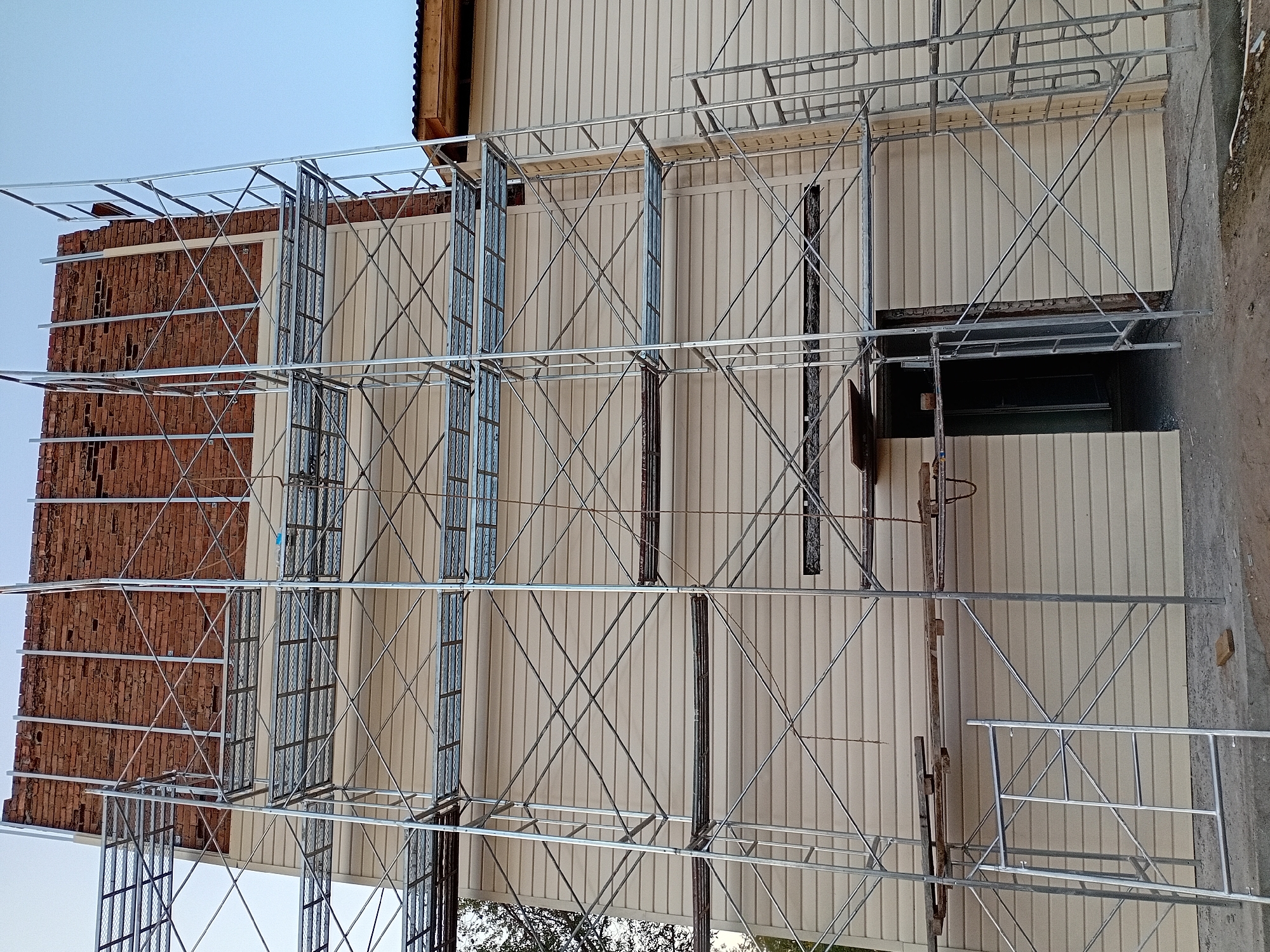 The image depicts a building under construction, possibly in the city of Semey, Kazakhstan, as indicated by the provided prompt. A large metal scaffolding structure is erected against the side of the building. The building features beige horizontal siding on its lower floors and exposed brickwork on its upper section, suggesting the final stages of siding installation or renovation. Several levels of scaffolding platforms are visible, with some wooden planks placed on one of the lower platforms, possibly for material storage or work surfaces. A dark opening, potentially a doorway or window, is visible in the siding on the ground floor, also surrounded by scaffolding. The sky is clear and blue, indicating daytime. The ground around the building appears to be dirt and gravel, consistent with a construction site. There are no people visible in the image.