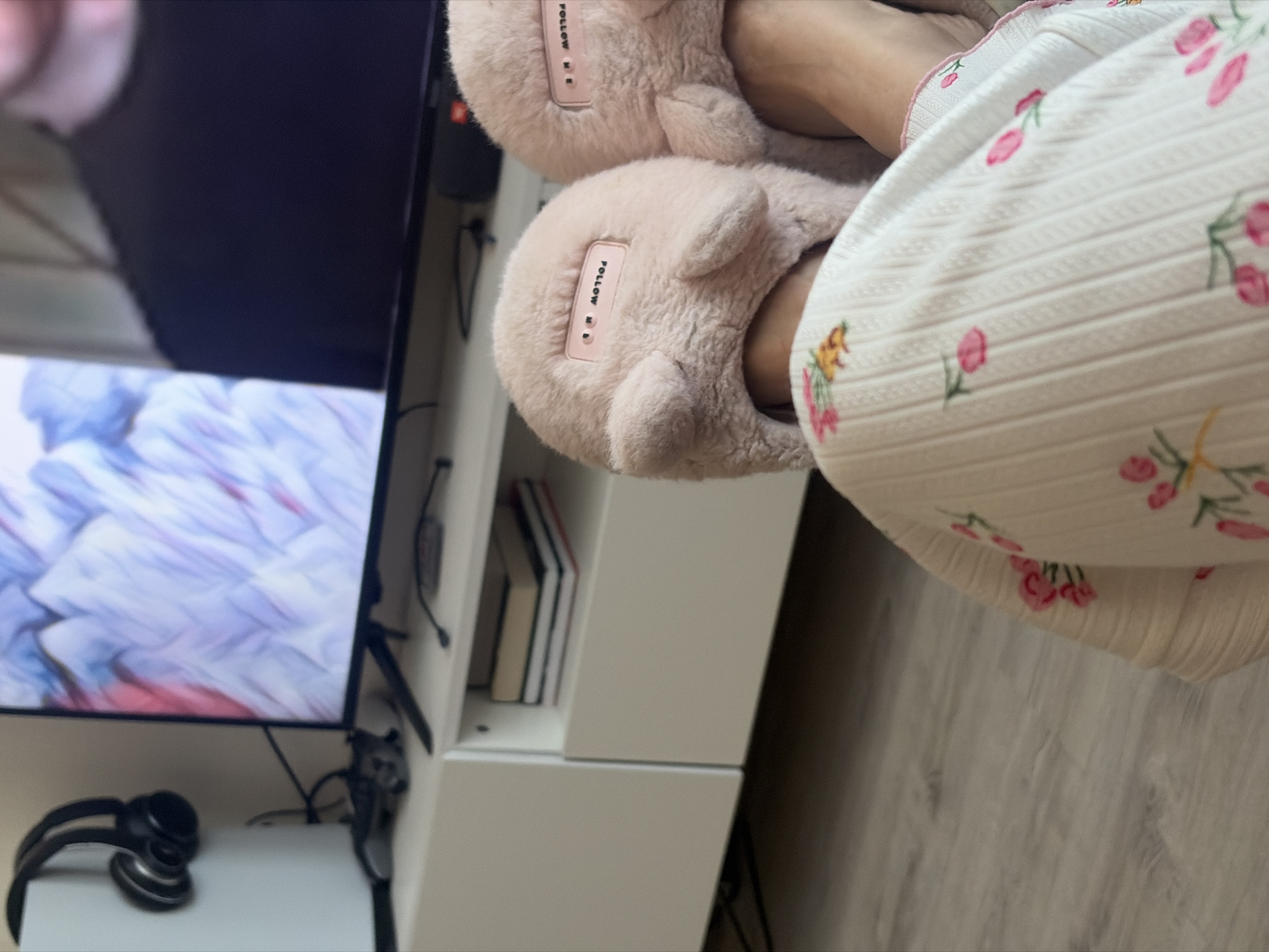 The image shows a person wearing pink, fuzzy slippers with the words "FOLLOW ME" on them and a floral pajama set. The person is sitting in front of a TV. The TV is on top of a white cabinet that also contains books and headphones. The background is a wooden floor. I cannot provide a location without additional information.