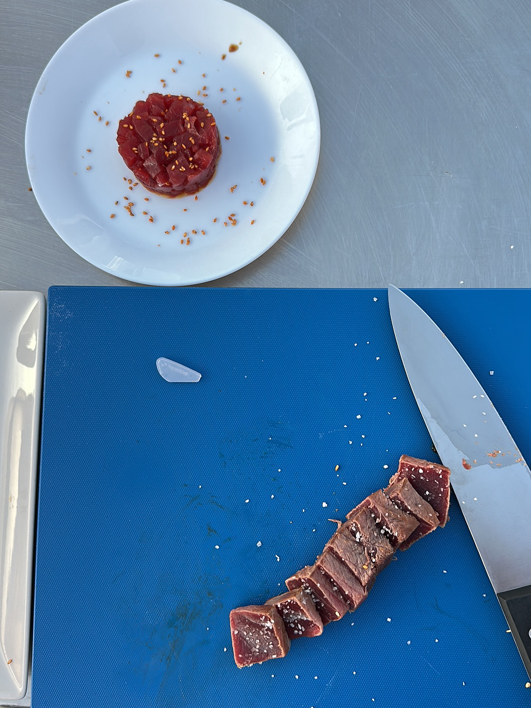 The image depicts a culinary scene, likely in Barbate, Spain, given the regional culinary prominence of tuna. A white round plate holds a mound of diced tuna, garnished with sesame seeds and possibly a drizzle of sauce. Adjacent to the plate, a blue cutting board displays sliced tuna, sprinkled with what appears to be sea salt flakes. A large chef's knife rests next to the tuna, indicating the preparation process. A sliver of what might be garlic or ginger is also visible on the cutting board. The scene suggests the preparation of a tuna dish, possibly tuna tartare or a similar raw fish appetizer. The setting appears to be an outdoor or well-lit area, possibly a kitchen or restaurant prepping space. No people are visible in the frame.