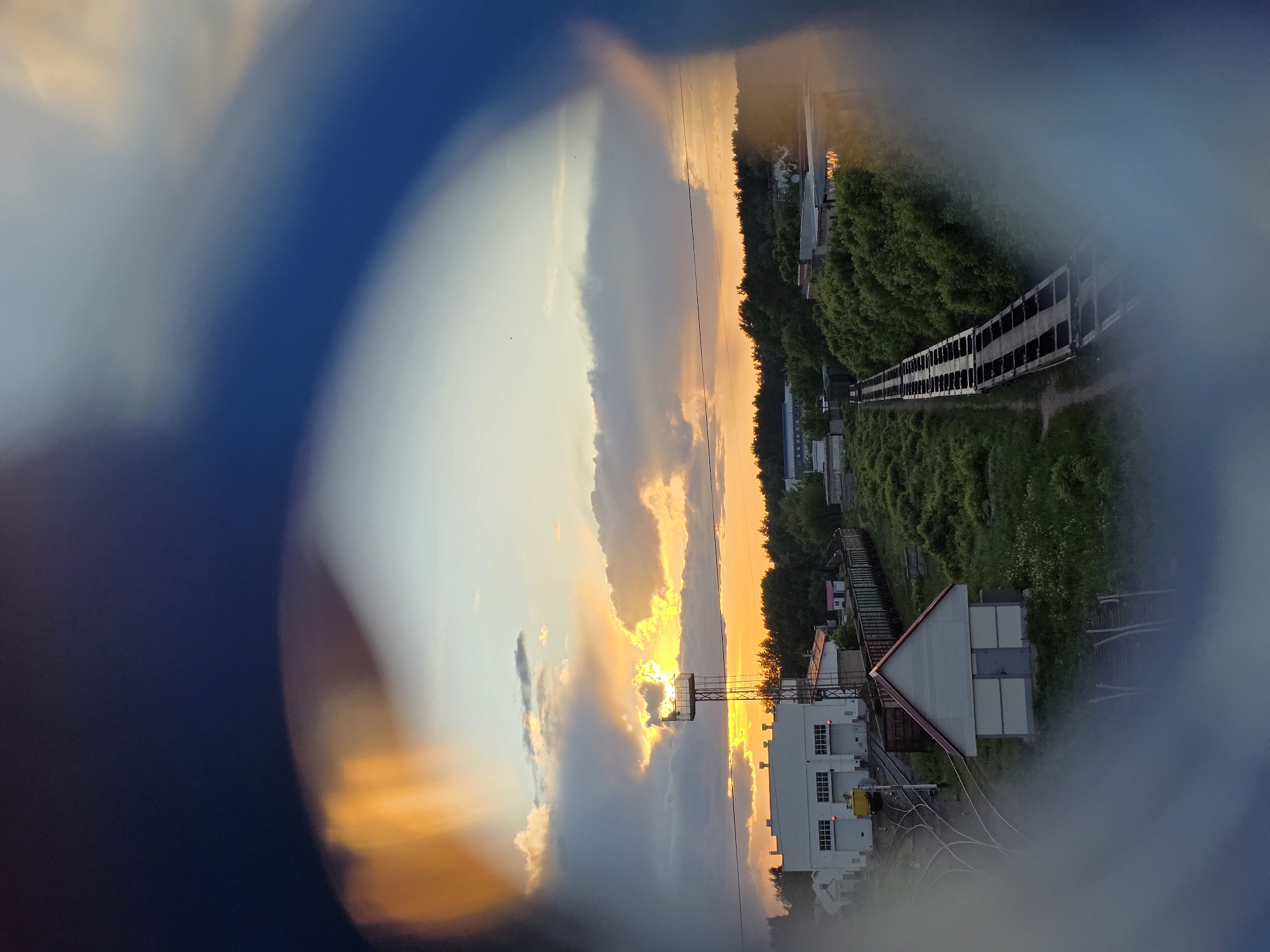 The image shows a landscape view through a circular lens. The lens is blurred around the edges, creating a vignette effect. The landscape includes a setting sun obscured by clouds, casting an orange glow. Below the horizon, there's a train bridge alongside greenery. There are a couple of buildings at the bottom, with one of them appearing white with windows. Some electrical wires run towards the structure. This image is most likely taken in the city of Chaikovsky, Russia based on the metadata.