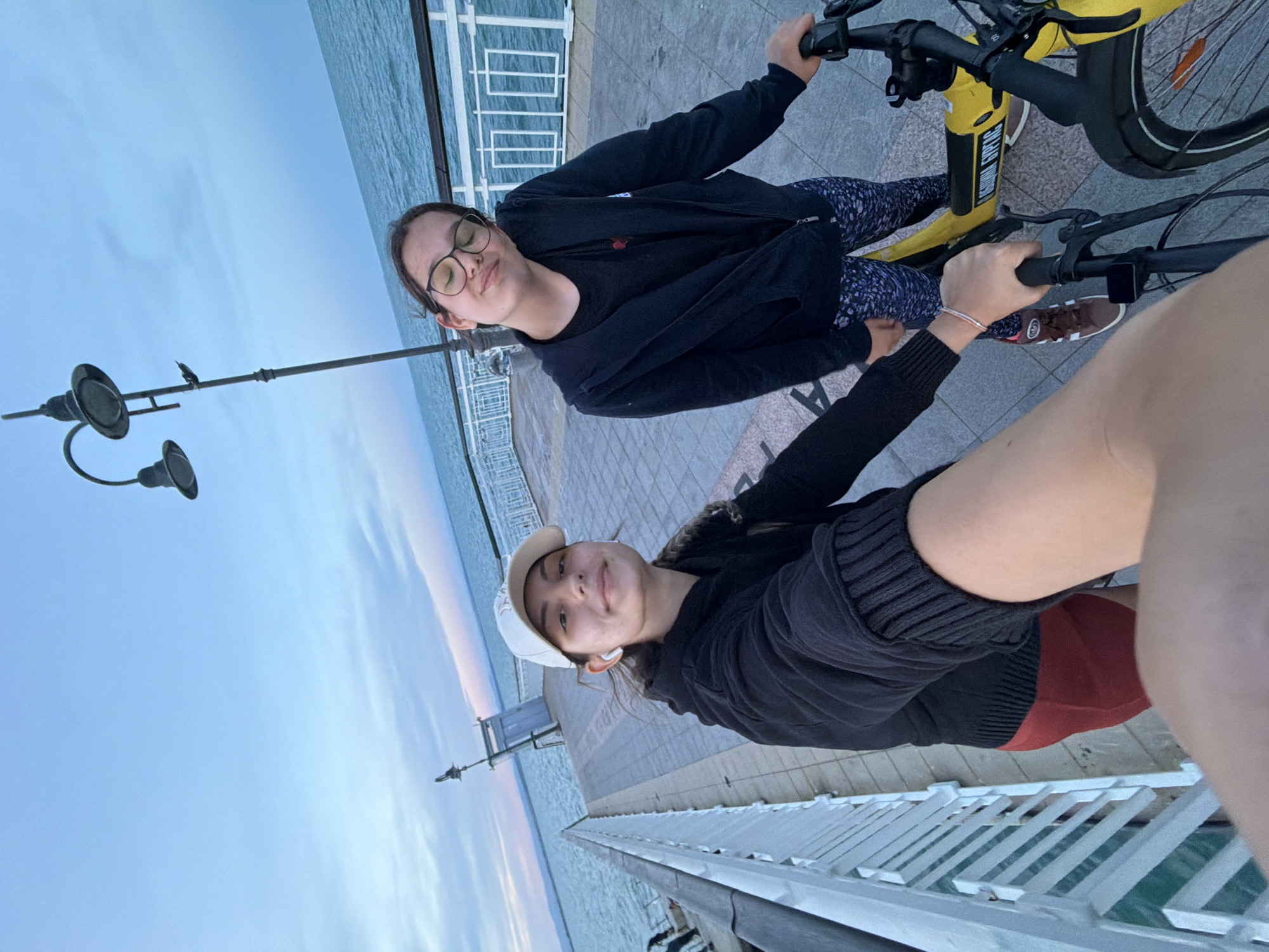Two young females are on a pier in Burgas, Bulgaria, with a body of water to their right and a cloudy sky above. One female, wearing a white baseball cap and black hoodie, is in the foreground, holding a bicycle handlebar. The other female, wearing glasses and a black hoodie, is behind her, also near a yellow bicycle. The pier has a paved walkway and a white railing. A lamppost is visible in the background. The overall lighting suggests it is either early morning or late evening.