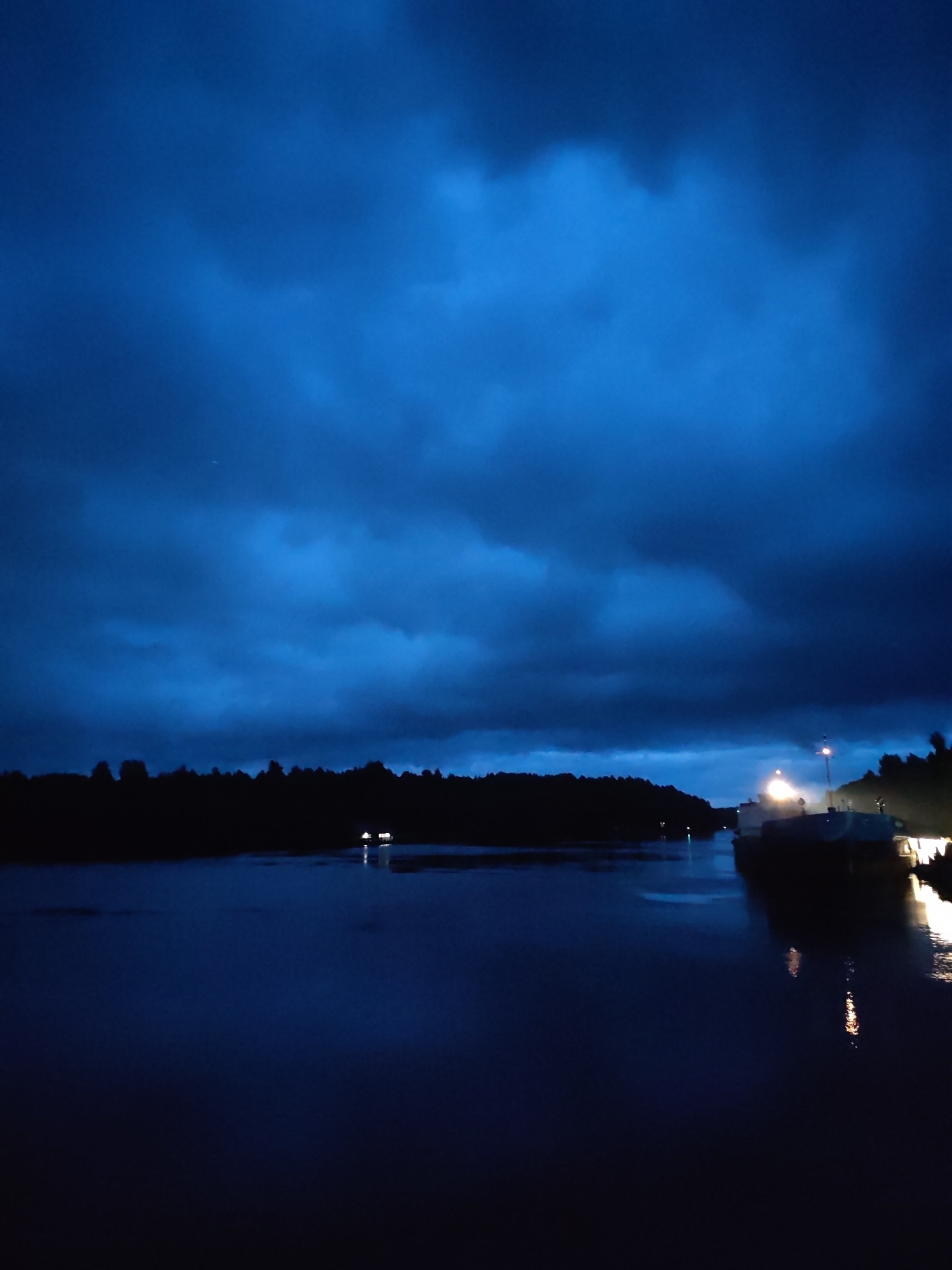 The image depicts a dark, overcast sky with deep blue hues and scattered clouds, suggesting either twilight or a storm approaching. Below the sky, a dark silhouette of a tree line runs across the horizon, separating the sky from a body of water in the foreground. The water is dark and reflects the dim light from the sky and some artificial lights. On the right side of the water, there are two boats docked. One boat is closer to the viewer and appears larger, with visible lighting. Another boat is further away, also illuminated. The scene is dark and quiet, with a sense of isolation. The location is identified as Devyatiny, Russia.