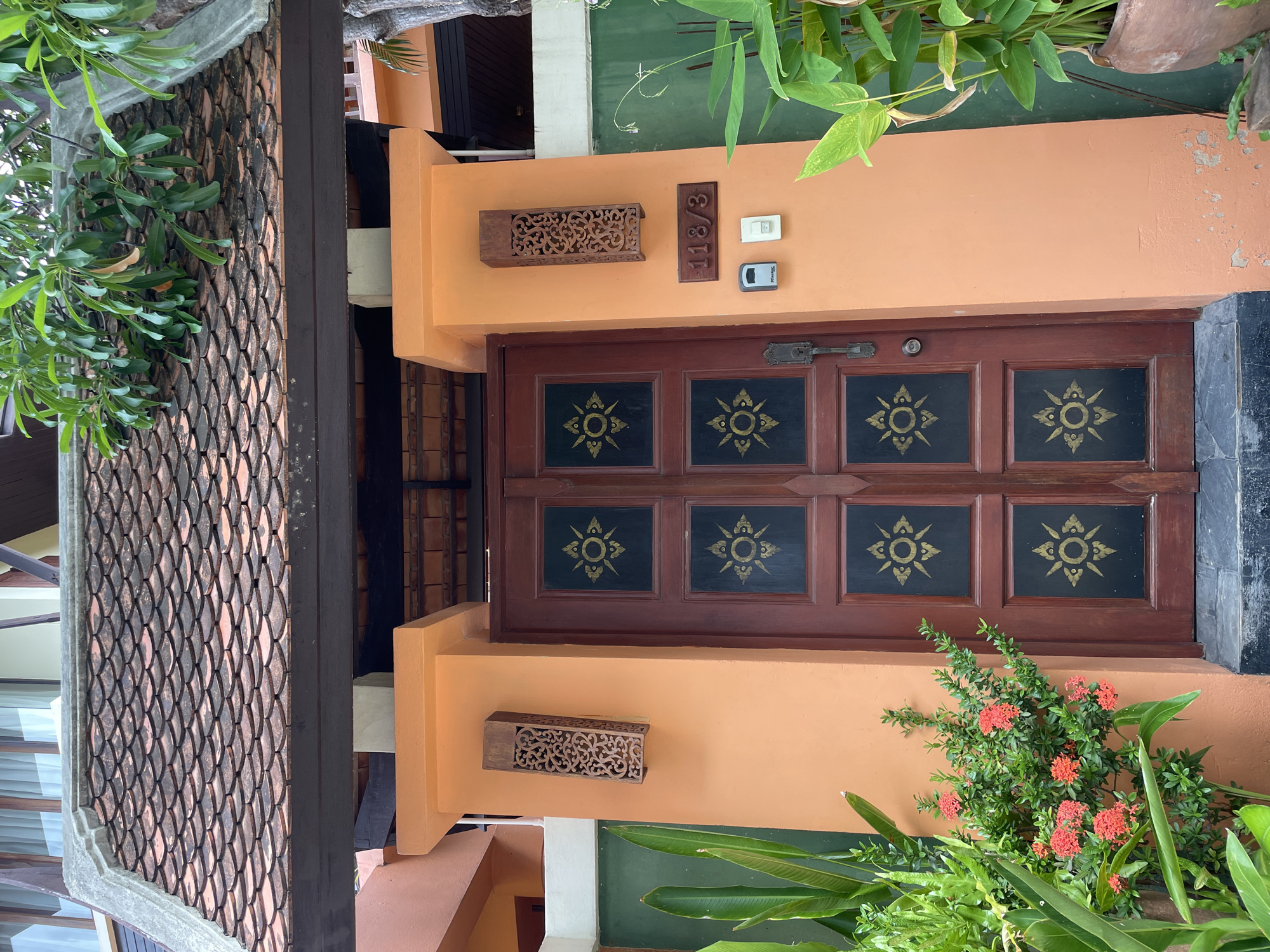 The image shows a closed wooden door with a dark frame and ornate gold designs on each of its six panels. The door is flanked by two orange pillars, each adorned with a carved wooden light fixture. Above the door is a tiled roof, and to the right, a dark wooden sign reads "118/3". Below the sign are two small electrical boxes. Lush green plants and a bush with small red flowers are visible on either side of the entrance, adding to the tropical atmosphere of the scene, likely located in Pattaya City, Thailand, judging by the architectural style and vegetation. The overall impression is one of a residential entrance with a distinctly Southeast Asian aesthetic.
