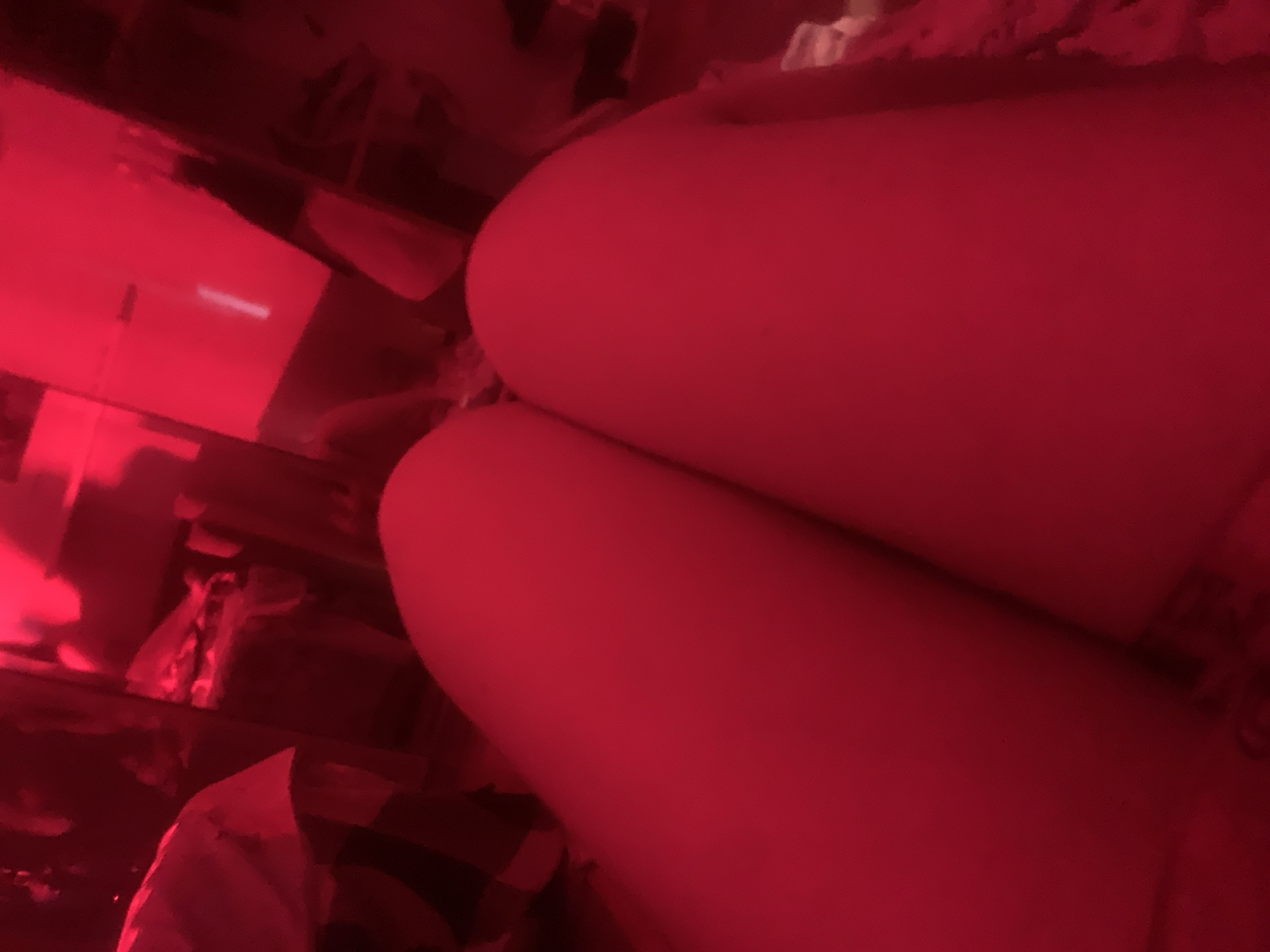 The image captures a person's lower body, specifically their upper legs or thighs, prominently displayed in the foreground. The individual appears to be in a relaxed pose, possibly sitting or lying down. Part of a patterned fabric, resembling plaid or check, is visible in the lower left of the frame, suggesting clothing or a blanket.
The scene is an indoor setting, likely a room, entirely bathed in a strong red light. This red illumination creates a dramatic and somewhat obscure atmosphere, making it difficult to discern specific details of the background, which contains indistinct objects that might be furniture or shelves.
No particular activity or event is clearly taking place beyond the person's posture. The notable detail is the pervasive red lighting, which obscures colors and fine textures, creating a mood that could be perceived as intimate, artistic, or mysterious. On a rectangular object in the mid-ground on the left, a faint, glowing horizontal line is visible, possibly from an electronic display or a light source, but no legible text can be identified. The time of day is not discernible due to the artificial lighting, but the ambiance suggests an evening or nighttime setting. The image itself does not contain any visual cues to confirm the location in Russia.