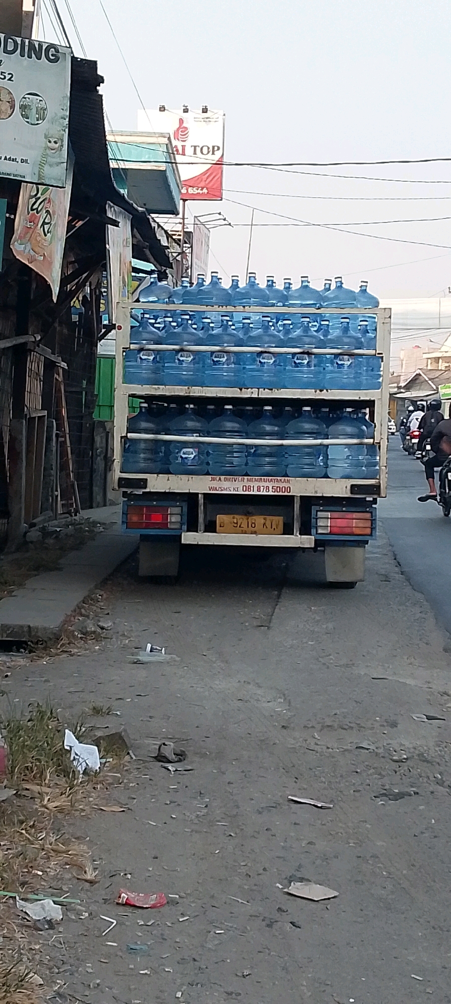 A delivery truck laden with large blue plastic water jugs is parked on a paved road in Karangsatria, Indonesia. The truck is filled with jugs stacked on two levels. The rear of the truck displays a license plate that reads "B 9218 XYV" and some text in Indonesian that translates to "IF THE DRIVER IS DANGEROUS, WHATSAPP TO 081 878 5000". In the background, there are buildings with signage, including one that says "AI TOP" with a telephone number and another that is partially obscured but appears to be an advertisement. Several people on motorcycles are visible further down the road, suggesting traffic. The immediate foreground and roadside are littered with various pieces of trash, including plastic wrappers, cardboard, and other debris. The sky is overcast.