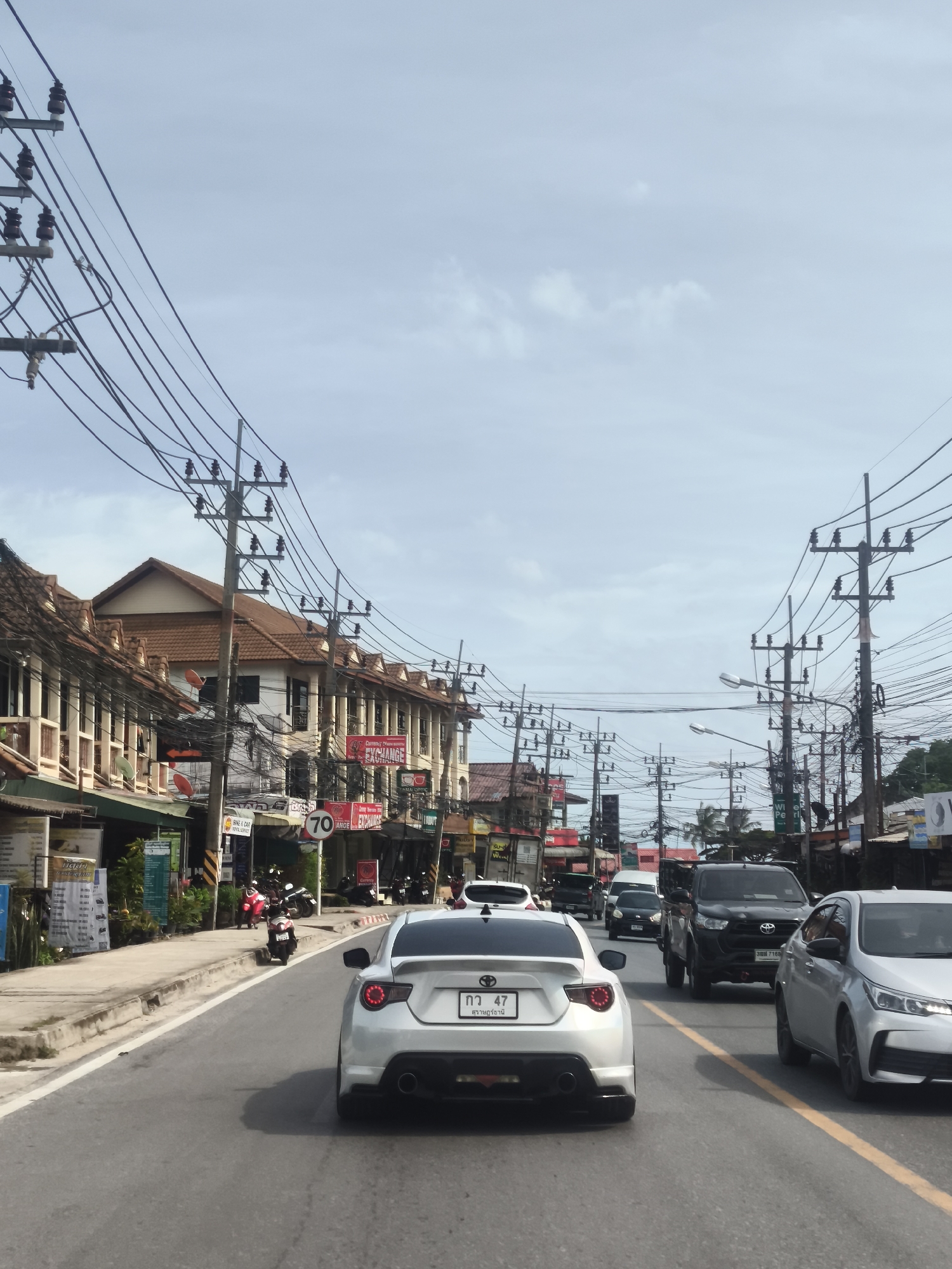 In a street scene on Ko Samui District, Thailand, traffic moves along a paved road lined with multi-story buildings housing shops and businesses. Utility poles with numerous wires are prominent along the roadside. A white sports car is in the foreground, followed by a silver sedan, and then a black pickup truck. Other vehicles, including motorcycles, are visible further down the road. The sky is overcast with some scattered clouds. A speed limit sign indicating "70" is mounted on a pole. The architecture of the buildings suggests a tropical environment.