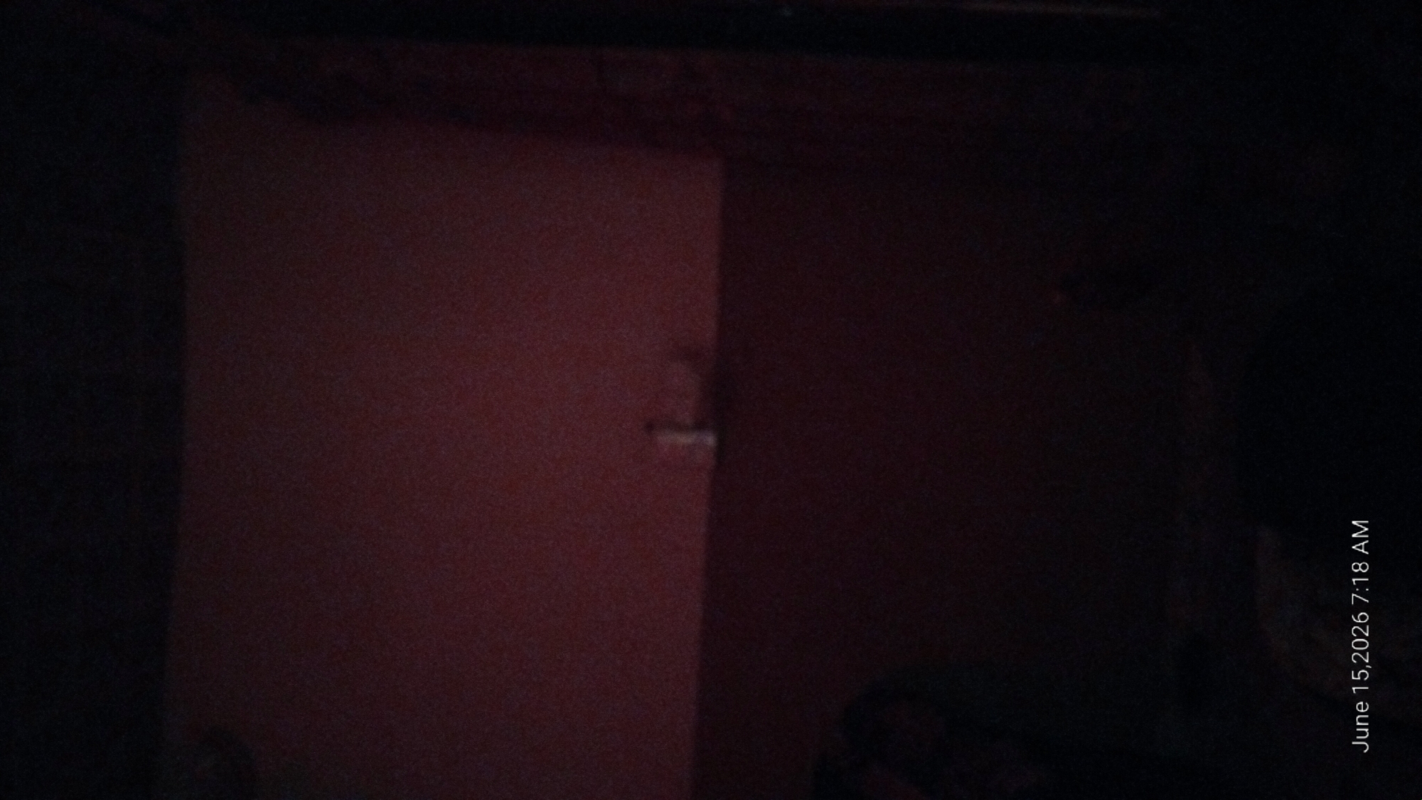 The image depicts a dimly lit interior scene. A horizontal line divides the frame, with the upper portion appearing to be a reddish-brown wall and the lower portion a darker, more indistinct area. A small, pale object, possibly a bottle or container, is visible on the dividing line towards the center of the frame. The bottom left of the image shows a dark, blurry shape that could be a person or furniture. The bottom right corner of the image contains a timestamp: "June 15, 2026 7:18 AM". There is no discernible information within the image that would allow for the identification of the location in Howayi, Nigeria.