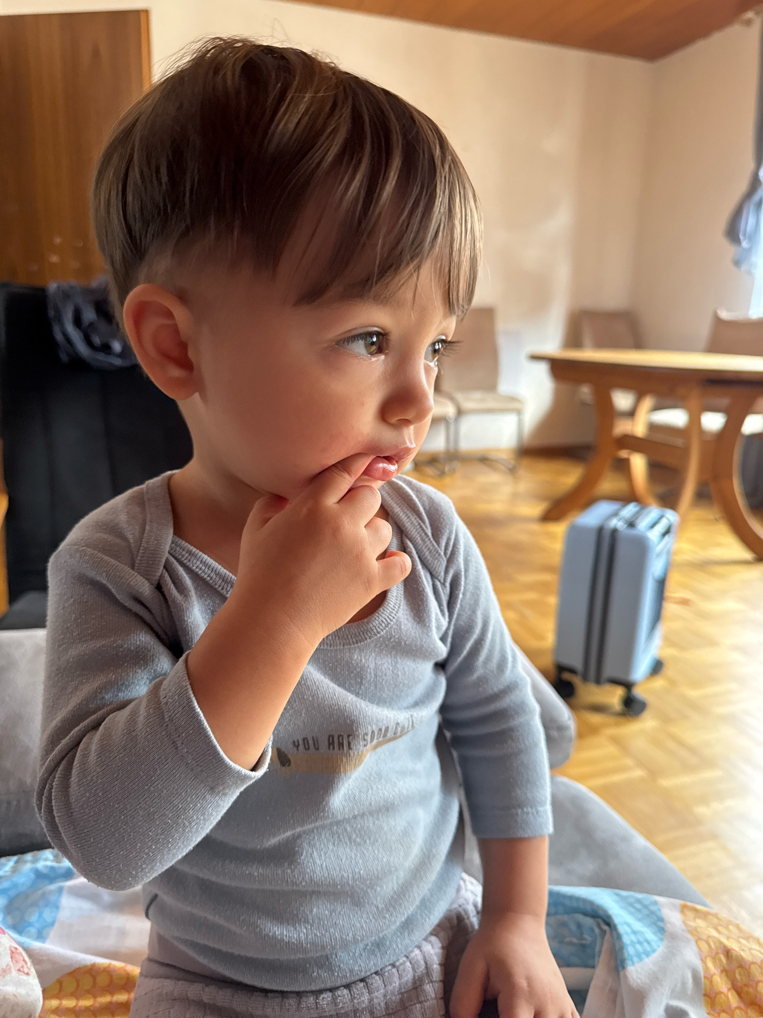The image shows a young child sitting and looking to the right, with their finger in their mouth. The child is wearing a light blue long-sleeved shirt and grey pants. In the background, there is a wooden table and chairs, a blue suitcase, and a wooden floor. The setting appears to be indoors, possibly a home in the city of Überherrn, Germany.