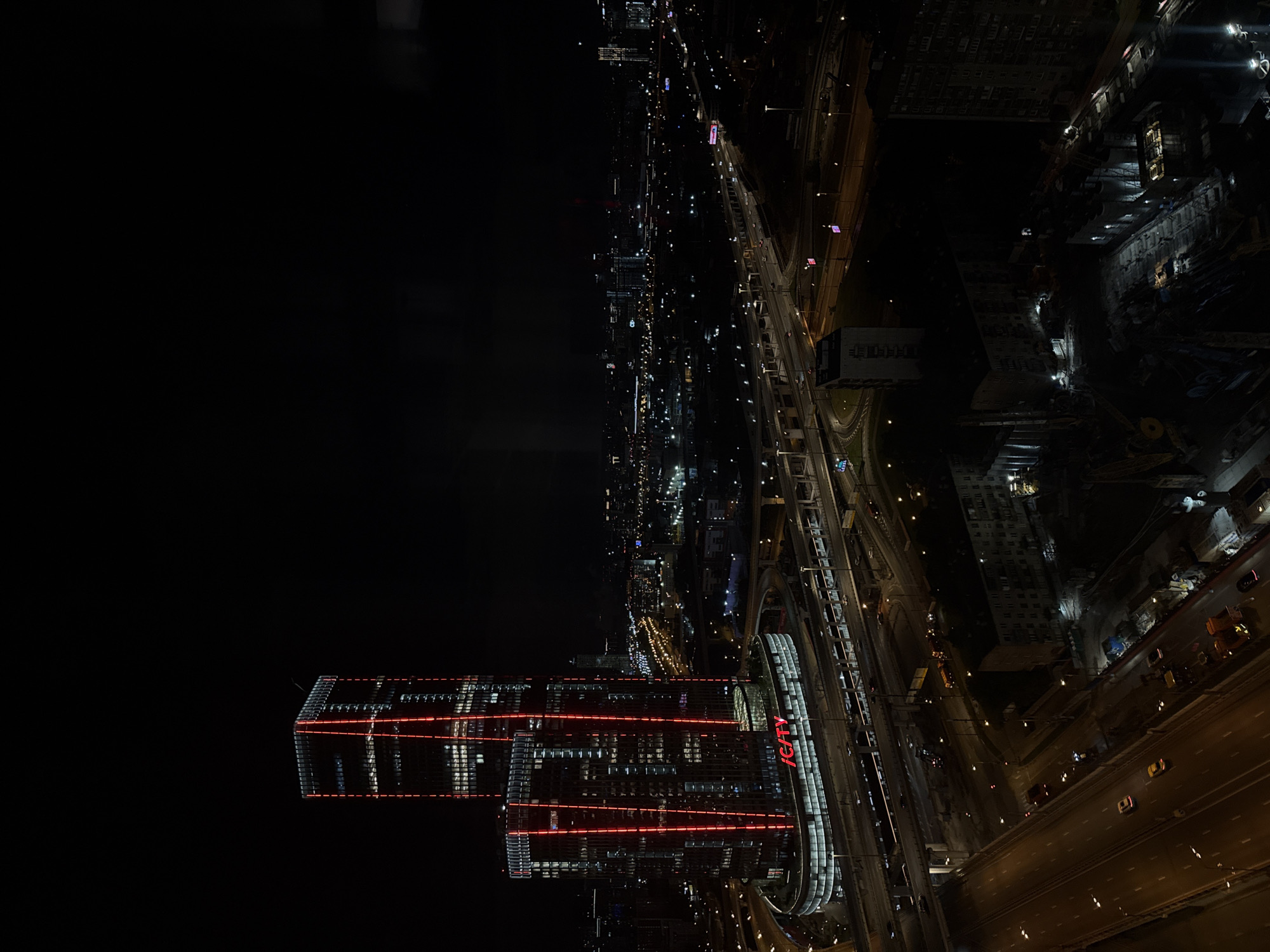 The image shows a nighttime aerial view of Khimki, Russia. Dominating the foreground are two modern high-rise buildings, illuminated with red horizontal lines. Adjacent to these buildings is a curved, illuminated structure with the letters "ICITY" vertically displayed. A multi-lane highway with moving vehicles stretches alongside the buildings. Further into the scene, the city's lights and buildings extend into the distance. The sky above is dark, providing a stark contrast to the city's lights.