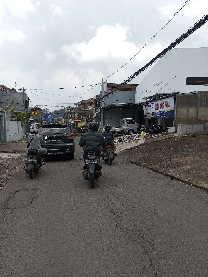The scene is located in Chendenbji, Bhutan, on an asphalt road under an overcast sky. The road surface exhibits cracks and patches, particularly in the foreground. Three motorcycles and one dark-colored SUV are present, all heading away from the viewer in a staggered formation.
From left to right: The first motorcycle is a grey scooter, ridden by an individual wearing a light grey hoodie, a white helmet with a dark visor, and a dark backpack. Its license plate is "F 2381 UAI". Directly ahead and slightly to the right of this motorcycle is a dark SUV with the license plate "F 1411 YJ". The second motorcycle, a dark grey scooter, is positioned center-right, operated by a person wearing a dark green puffer jacket and a dark helmet; its license plate reads "F 3432 YC". The third motorcycle, dark in color, is further right, with its rider wearing a dark jacket and helmet.
On the left side of the road, multiple multi-story buildings with varied facades and visible red-tiled roofs line the street. A dense network of overhead electrical and communication wires spans above the road. A partially visible yellow sign with a "CP" logo is present on one building. Further down, the street appears to continue into a more densely built-up area.
On the right side of the road, a large, white-painted building dominates the mid-ground. Beneath it, an open-front structure houses a light-colored utility truck, possibly a small flatbed. A prominent blue and white banner is affixed to the white building, displaying the word "MAPAT" and images of various food items. Above this banner, a dark, elevated sign with red lettering is visible. The ground immediately to the right of the road is uneven and littered with debris, including a yellow plastic bag. Additional overhead wires extend across this side of the street.