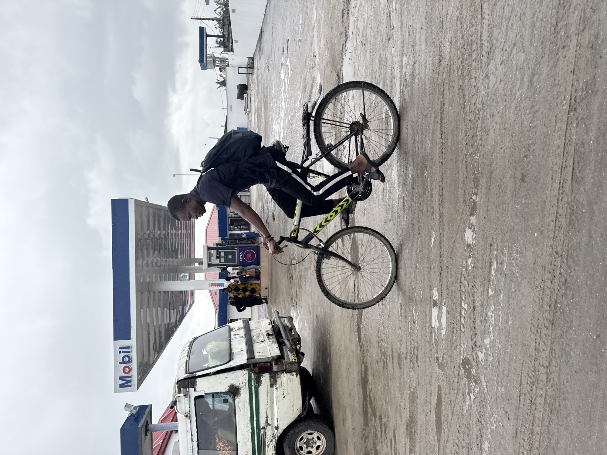 A young man wearing a dark shirt, dark pants with a white stripe, and black sandals rides a bicycle with a black frame, yellow accents, and a black backpack. He is at a gas station, identifiable by the white and blue "Mobil" signage and a white van parked to his left. The ground is wet and appears to be muddy asphalt. In the background, other people are visible near the gas pumps, and a white building with a pink roof is present. The sky is overcast. The scene appears to be a typical street or gas station environment in Ado, Nigeria.
