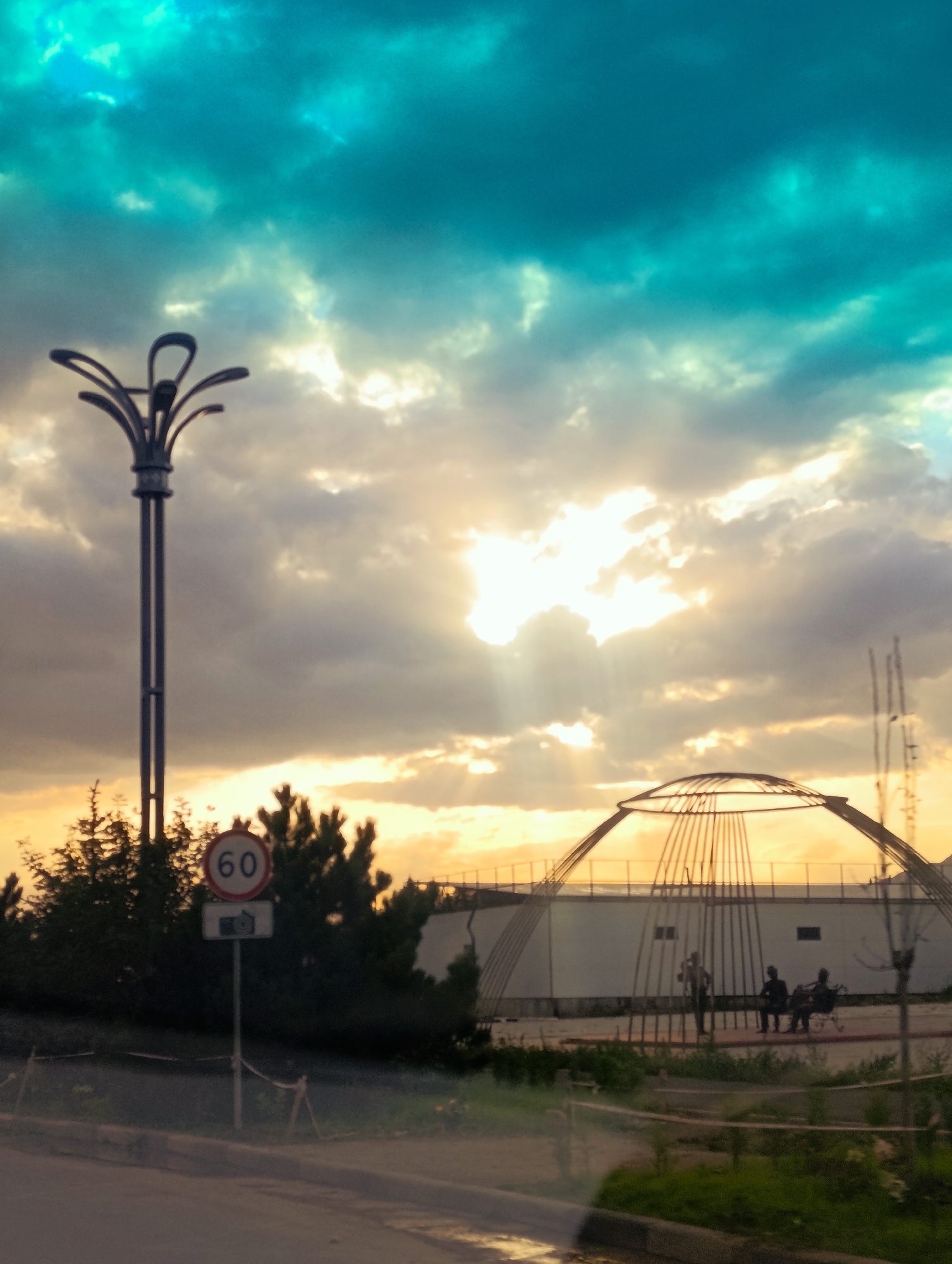 An eye-level, full shot captures a dramatic sunset sky over a street scene in Balykchy, Kyrgyzstan. The sky is a vibrant mix of turquoise and muted orange, with bright rays of sun piercing through thick, dark clouds. In the foreground, a dark lamppost with a decorative top stands to the left, and a speed limit sign displaying "60" is visible on a pole with a camera symbol below it. To the right, a modern, dome-like structure with an open framework is under construction or partially built. Silhouetted figures can be seen near the structure; one person stands, and two others are seated on a bench. A white building with dark windows is situated behind the dome. The ground in the foreground appears wet, suggesting recent rain. Greenery, including trees and grass, lines the edge of the paved area, and a road is visible in the lower portion of the frame.