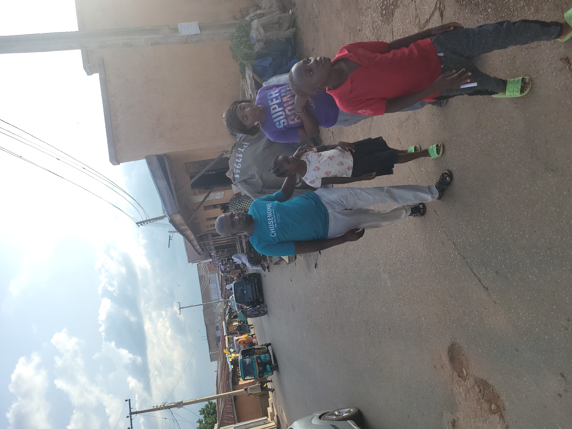 The image depicts an outdoor street scene in what is identified as a city in Jos, Nigeria, under a bright, partly cloudy sky. The primary subjects are a group of four individuals walking on an asphalt road in the foreground, moving towards the viewer's left.
From left to right:
1. A bald adult male in a teal t-shirt with "CHOSENONE" text and light-colored trousers, wearing dark sandals. His left arm is around the shoulders of a young girl. He is looking slightly to his left.
2. A young girl, a child, in a white short-sleeved top with a floral pattern and a black skirt, wearing light green slides. She is holding the man's left hand and looking straight ahead.
3. An adult female in a purple t-shirt with "SUPER BOWL" text and light grey trousers, wearing dark sandals. She is walking forward, looking slightly to her right.
4. A young boy, a child, in a red polo shirt and dark grey trousers, wearing light green slides. He is walking forward, looking to his right.
The road surface shows wear, with visible potholes in the mid-ground on the left side.
In the mid-ground, a green and yellow motorized tricycle (Keke Napep) with "PN 5591" visible on its side is driving away on the left lane. A dark-colored car is parked or slowly moving on the right lane further down the street. A light-colored car is partially visible on the far left edge of the frame.
Buildings line both sides of the street. These structures are predominantly single-story, with light brown or tan facades and a mix of flat roofs and corrugated iron roofs. Many appear to be commercial establishments or market stalls. On the right, a stall covered by tarpaulins displays green leafy produce. A white sign with "NGO" is affixed to a utility pole on the right.
Multiple wooden and concrete utility poles equipped with numerous overhead wires are visible throughout the scene, indicating electrical or communication infrastructure. The sky is bright with scattered cumulus clouds, and strong sunlight originates from the upper right, causing some lens flare in that area. Several indistinct figures are present in the background, walking or standing near the buildings.