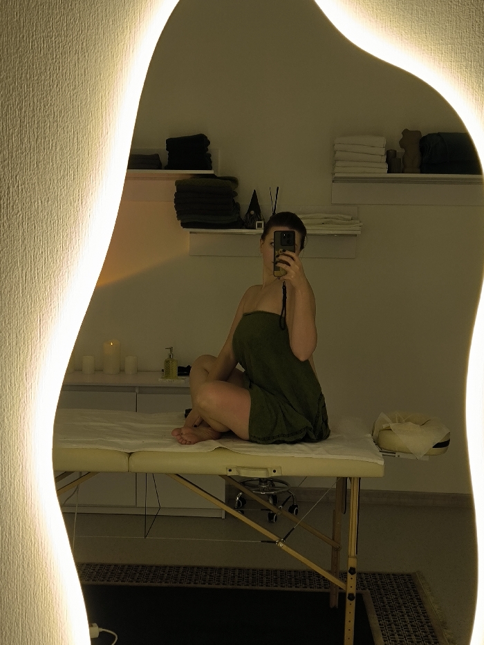This image is a reflection in an irregularly shaped, backlit mirror, depicting an indoor scene. A single female subject is seated cross-legged on a beige portable massage table, which is covered with a white sheet. She is wrapped in a dark olive-green towel, exposing her bare feet. The subject is holding a smartphone with a yellow case in her right hand, pointed towards the mirror, indicating she is taking a selfie.
In the background, multiple white shelves are mounted on a light-colored wall. These shelves display stacks of neatly folded dark and white towels, a small dark gnome figurine, and at least two reed diffusers. To the left, on a white counter, a tall lit candle, two smaller white candles, and a clear pump dispenser containing a yellow liquid are visible. The room has soft, ambient lighting. The floor beneath the massage table features a dark textured mat. The specific location within Vinnytsia, Ukraine, cannot be determined from the visual information provided in the image.