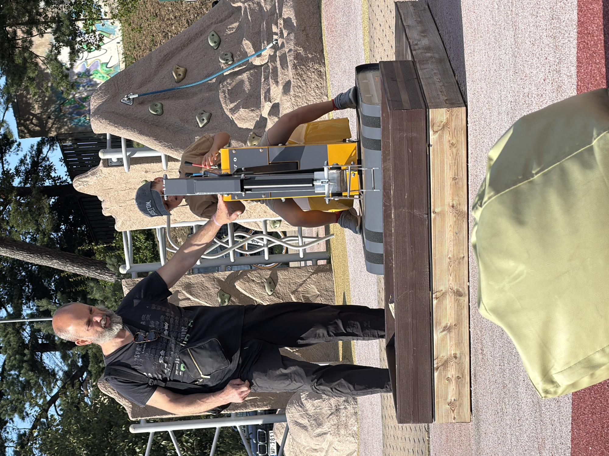 The image depicts an outdoor scene at a playground in Ventspils, Latvia. A man with a beard and a black t-shirt is standing on a wooden platform, looking down and gesturing towards a child. The child, wearing a beige t-shirt and a baseball cap, is seated on a yellow and gray mechanical ride or toy. The playground features a climbing wall with colorful holds in the background, as well as trees and a fence. The ground is covered in a patterned surface of red and beige material. In the foreground, a green beanbag chair is partially visible. A car with license plate "MP-7813" is parked to the left of the frame. The lighting suggests it is daytime, with shadows cast by the structures and trees.