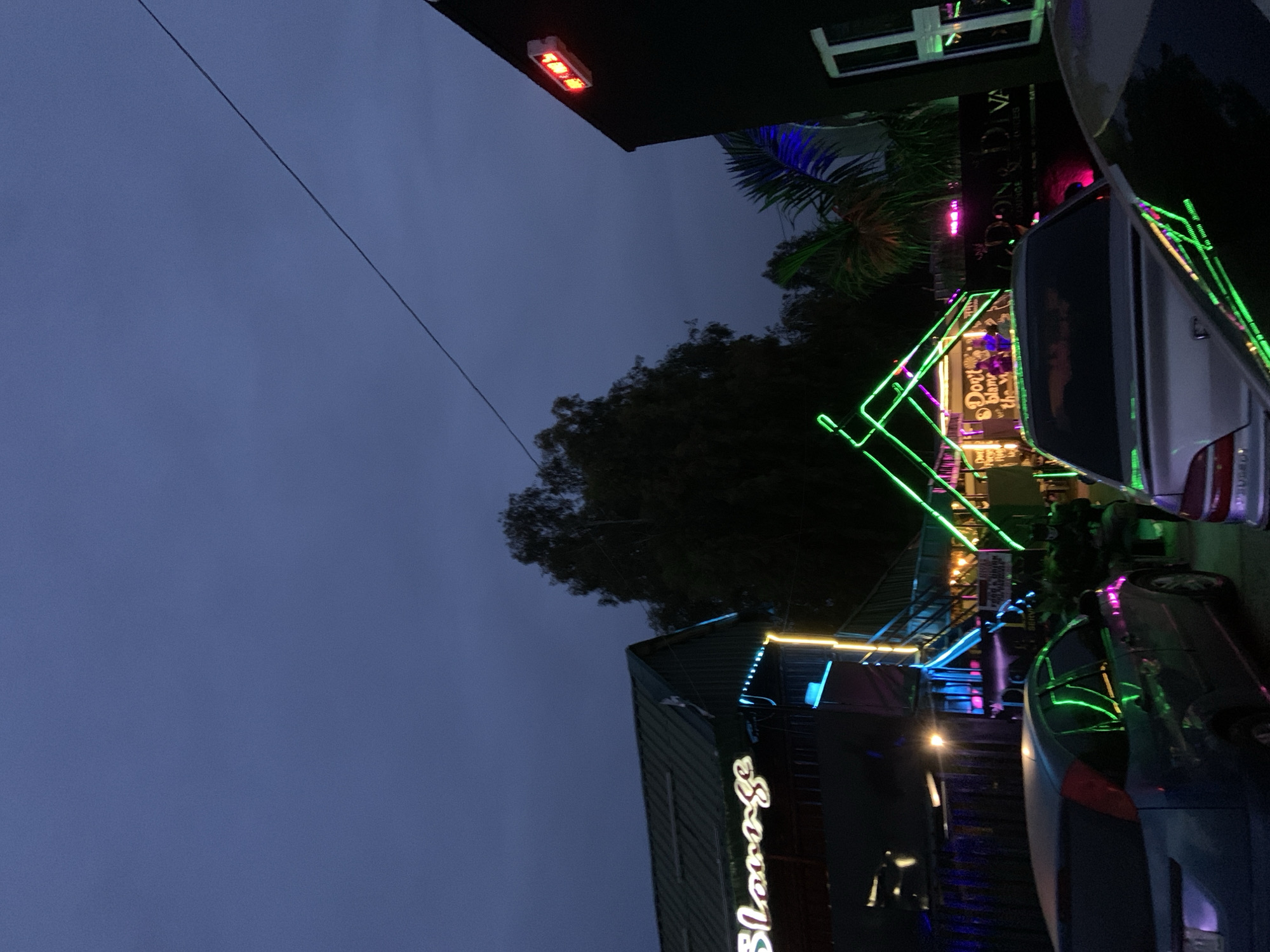 The image depicts an outdoor scene at dusk, possibly in the city of Gura Top, Nigeria, characterized by vibrant neon lights and several vehicles. In the foreground, two cars are visible, one silver sedan and a darker colored hatchback, parked in a lot. Behind the cars, a structure with green and yellow neon lighting is prominent, suggesting it's a business or entertainment venue. The signage on this structure appears to read "Slorge" in neon letters. Further back, another building is visible with a red light fixture on its facade. Trees and foliage are present in the background, creating a natural element against the artificial lights. The overall atmosphere suggests a lively evening setting.