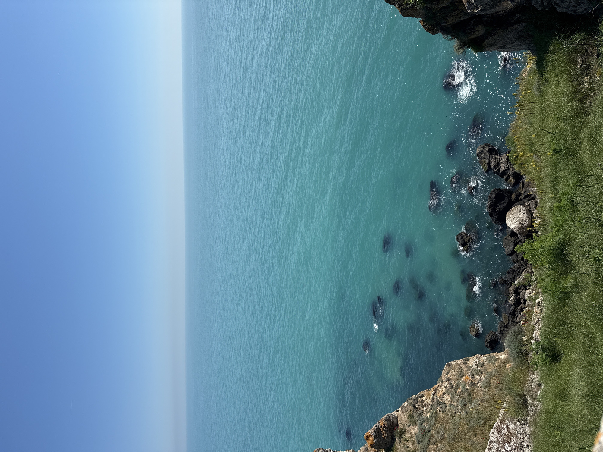 A vertical aerial shot depicts the coastal landscape near Balgarevo, Bulgaria. The deep blue sky occupies the left portion of the frame, transitioning to a lighter turquoise-colored sea. The sea's surface has slight ripples. The right edge of the image shows a rocky coastline covered in green vegetation. Several dark rocks are visible in the water close to the coastline. The cliff face is visible in the bottom portion of the image.