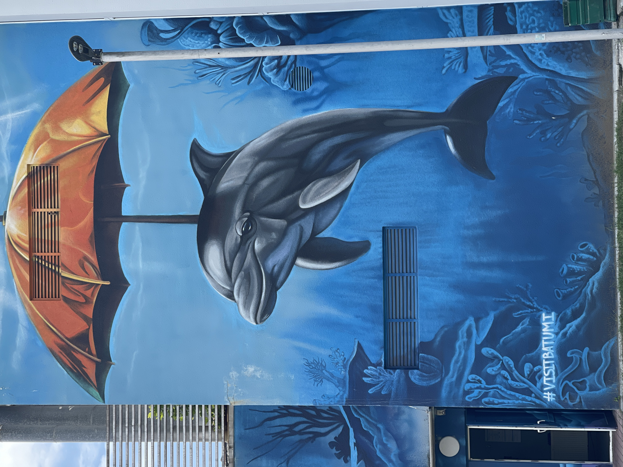 This is a vertical, close-up photograph of a mural painted on the side of a building. The mural depicts an underwater scene in shades of blue, with a gray dolphin prominently featured swimming towards the viewer. To the left of the dolphin is an open, orange umbrella, positioned as if it is part of the underwater environment. Various coral and plant-like forms are painted throughout the blue background, suggesting a marine setting.
Two grey rectangular grates are integrated into the mural, one to the left of the dolphin, appearing to be part of the umbrella's structure, and another to the right of the dolphin. A street lamp is mounted on a silver pole that extends horizontally across the top portion of the mural. The mural appears to be on an exterior wall, and in the bottom right corner, the hashtag "#VISITBATUMI" is painted in white. The overall impression is artistic and imaginative, blending terrestrial and aquatic elements.