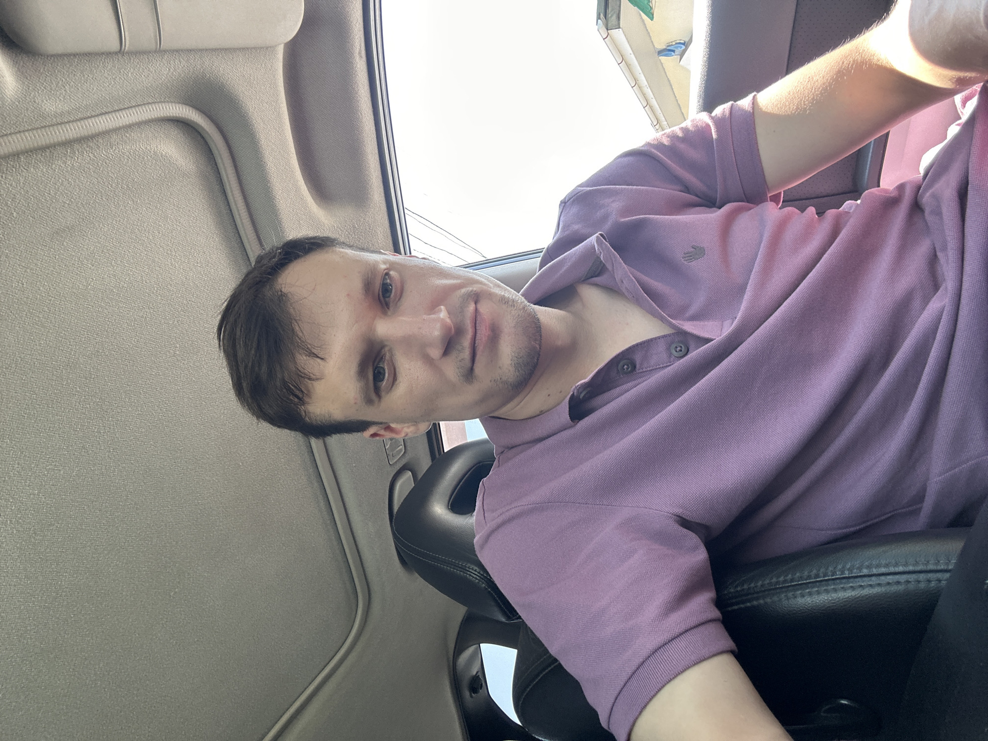 A man with short brown hair and blue eyes is sitting in the driver's seat of a car. He is wearing a light purple polo shirt. The car has a sunroof, which is open. The interior of the car is dark leather. The man is looking at the camera and smiling slightly. Outside the car, it appears to be a sunny day. In the background, there are some buildings and trees. The location is the city of Kamianets-Podilskyi, Ukraine.
