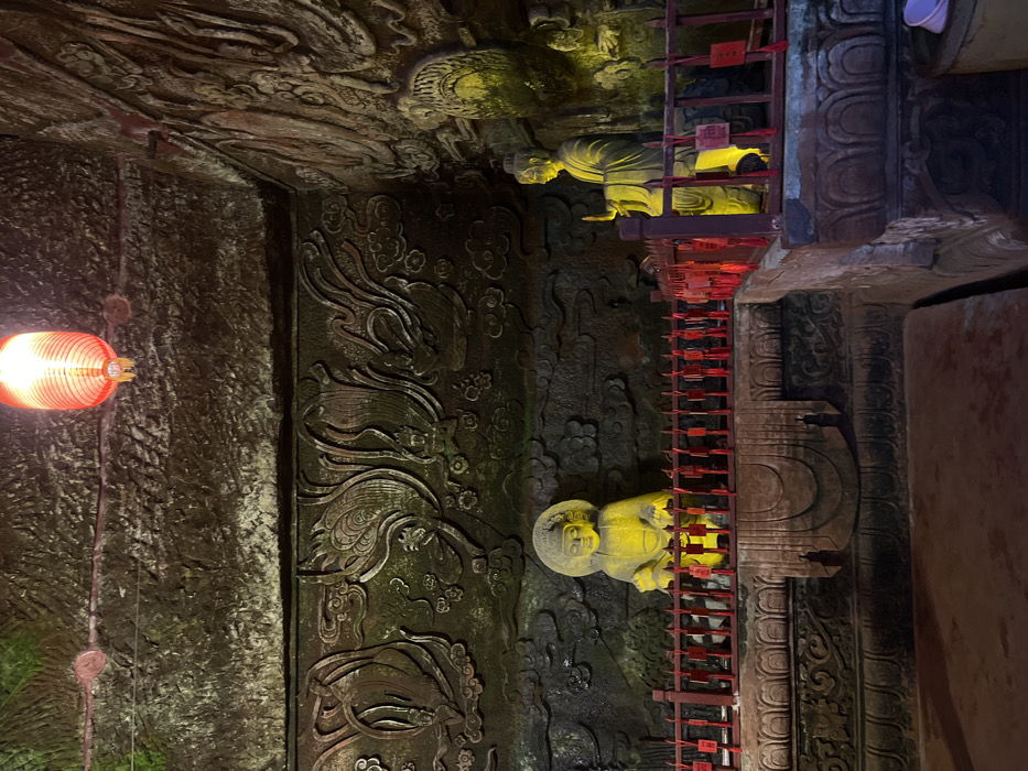 This image depicts the interior of a stone grotto or temple, specifically located in Shizhong, China. The scene is illuminated by artificial light sources, creating areas of yellow and green light against predominantly dark stone surfaces.
The ceiling is a rough, unworked stone surface, from which a single red, ribbed Chinese lantern is suspended towards the upper center, emitting a warm glow.
The walls are extensively carved with intricate bas-reliefs depicting figures, possibly celestial beings or deities, interspersed with swirling cloud motifs and decorative patterns. These carvings cover the visible back wall and extend around the corner to the right. The stone appears dark and textured, with some areas exhibiting green discoloration, possibly moss or mineral stains, particularly noticeable under the illumination.
Two primary stone statues are visible. On the left, a seated Buddha figure, characterized by a halo-like element behind its head, is brightly illuminated in yellow light. To its right, a standing figure, likely a Bodhisattva, with its right hand raised, is also illuminated in yellow light. Both figures are situated on elevated, carved stone platforms that feature decorative lotus petal designs.
A red wooden railing runs horizontally across the scene, positioned in front of both statues. Numerous small, rectangular red tags, presumably prayer or wish plaques, are tied to this railing with red strings.
The floor in the foreground is a dark, unadorned stone surface. In the lower right corner, a light-colored, possibly purple, plastic container is visible on the floor.