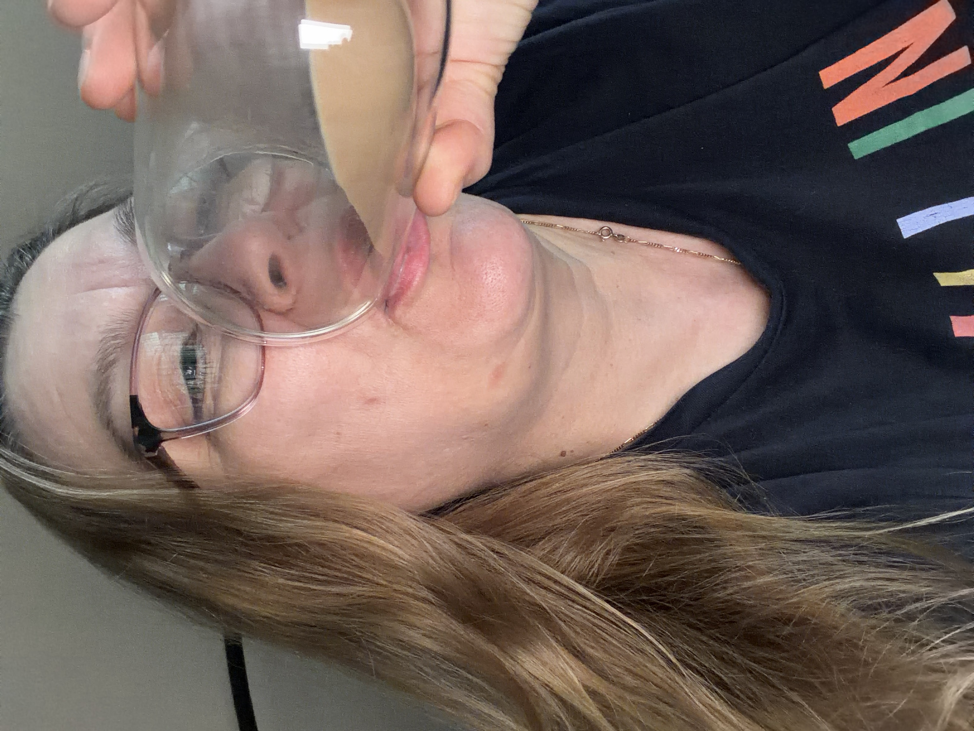 A woman is shown from the chest up, wearing glasses and drinking from a glass. She has long, blonde hair and is wearing a black t-shirt with colorful lettering. The background is a plain, light gray wall. The image is a close-up, with the woman's face and upper body filling the frame. The scene appears to be indoors, in a domestic setting. There is no indication of the location being in the city of Southend-on-Sea, United Kingdom.
