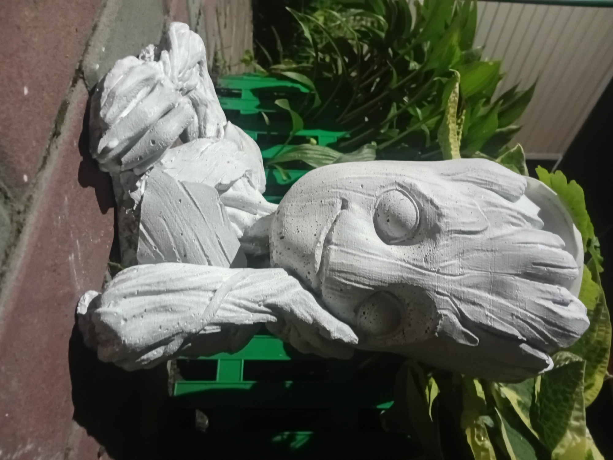 A grey, concrete statue resembling the character Groot from Guardians of the Galaxy is positioned outdoors on a brick-like pathway. The statue depicts Groot's upper body and head, with its right hand resting against its cheek and its left arm outstretched, resting on the ground. The statue is white and textured, with visible cracks and imperfections. Behind the statue, lush green foliage is visible, with some leaves featuring variegated patterns of green and yellow. A bright green fence runs horizontally in the background, partially obscured by the plants. The lighting suggests it is nighttime or dusk, with a spotlight illuminating the statue from the front, casting a distinct shadow to the right. The overall scene appears to be a garden or outdoor decorative area.