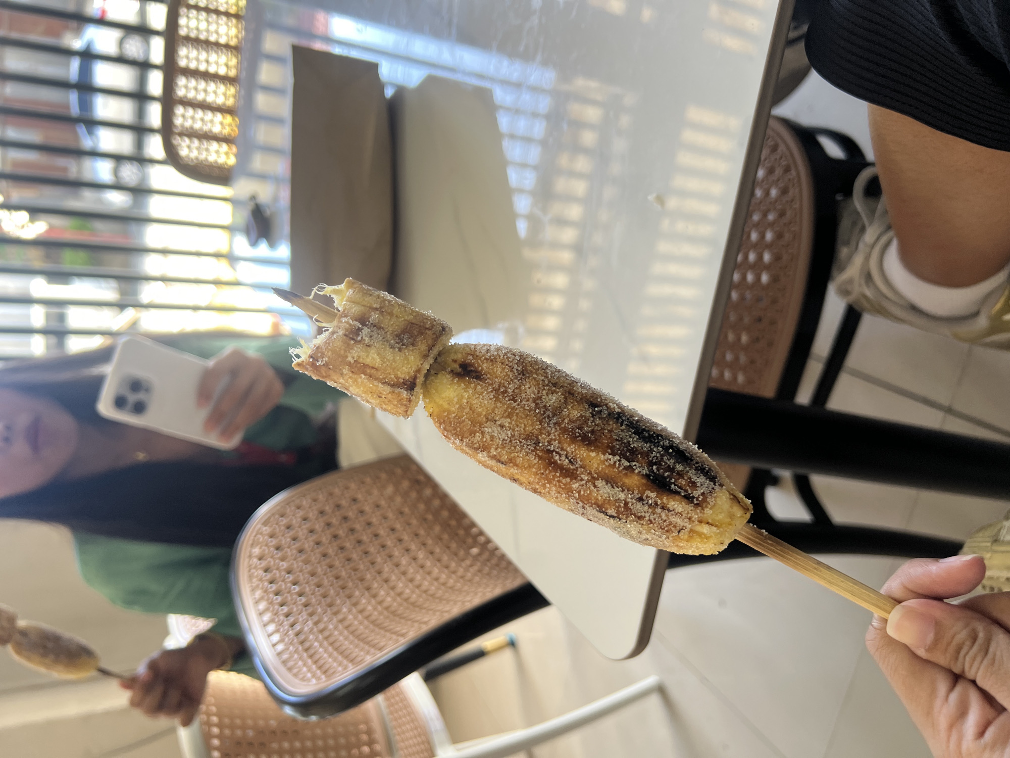 A person is holding a grilled banana skewer, heavily coated with sugar, at a table inside what appears to be a food establishment. A second person can be seen in the reflection in the table holding a mobile phone, possibly taking a picture of the banana. Wicker chairs are visible, indicating a casual dining atmosphere. The scene is brightly lit, possibly during daytime. The location is likely Davao City, Philippines, as indicated in the prompt.