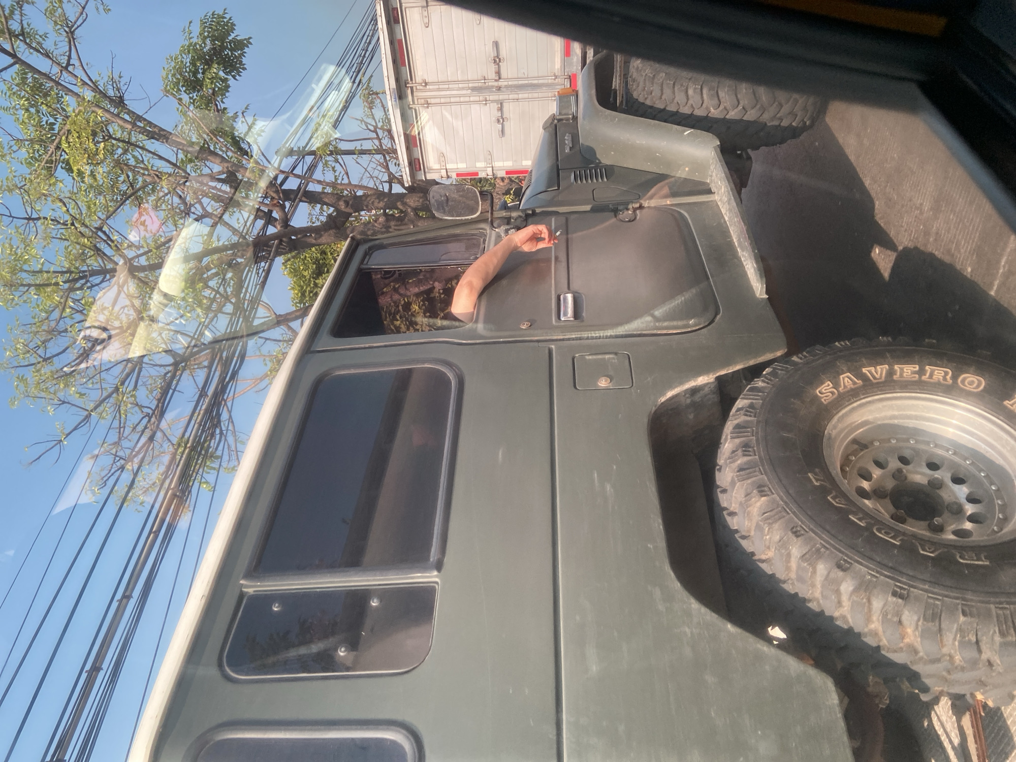 The image, captured on a sunny day in Semarang, Indonesia, shows the side profile of a dark green, vintage SUV parked on a street. The SUV has large, off-road tires and tinted windows. A person's arm is visible resting on the open driver's side window, holding a lit cigarette. Behind the SUV, a white semi-trailer truck with closed doors is also visible. Overhead, power lines and a leafy tree with green foliage are present against a clear blue sky. The sunlight creates reflections on the SUV's windows and body. The scene appears to be a street in a city, possibly during traffic.