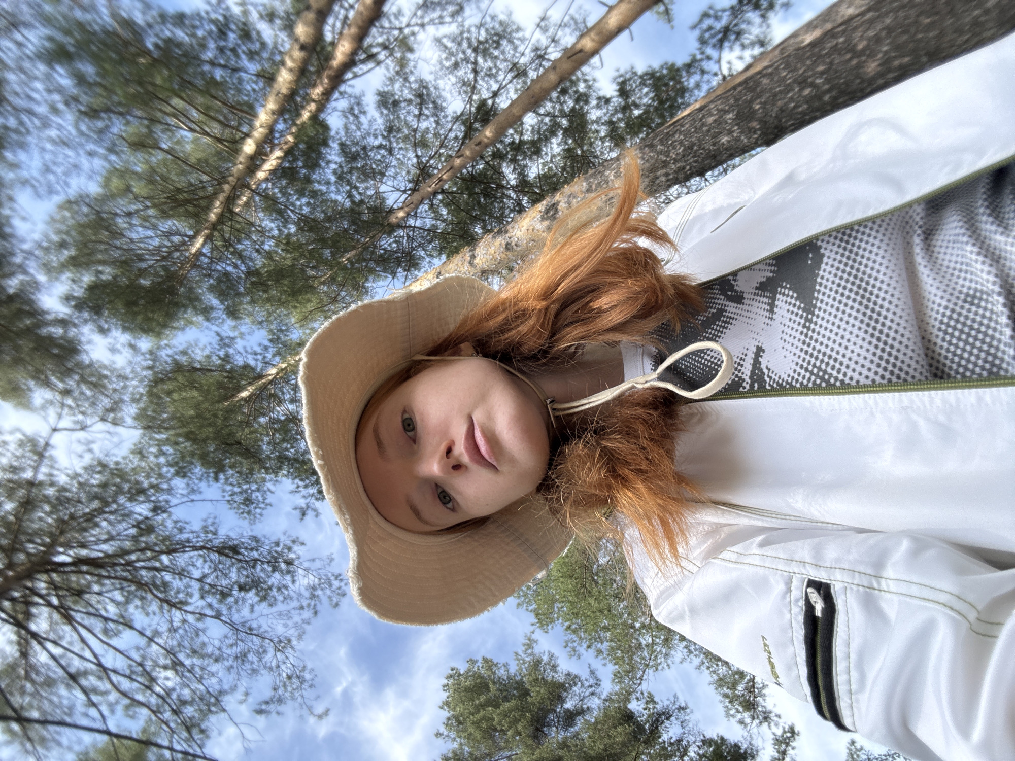 An image, presented rotated 90 degrees counter-clockwise, shows a young individual outdoors in what appears to be a pine forest, likely in Chaikovsky, Russia. The person, with fair complexion, light-colored eyes, and prominent reddish-brown hair, looks directly at the camera with a neutral to slightly contemplative expression. They are wearing a light-colored, possibly beige or off-white, wide-brimmed bucket hat secured by a chin strap. Over a grey, black, and white patterned top, they don a white jacket or windbreaker with olive green trim. A zippered pocket is visible on the jacket's left sleeve, above which faint, partially obscured text, possibly "VRSHOW" or "VRESHON", can be seen. The background features tall pine trees with rough, light-colored bark and green needles, viewed from a low angle against a clear blue sky with scattered white clouds, indicating a bright daytime setting. The scene suggests an outdoor activity such as a walk or hike in a forest.