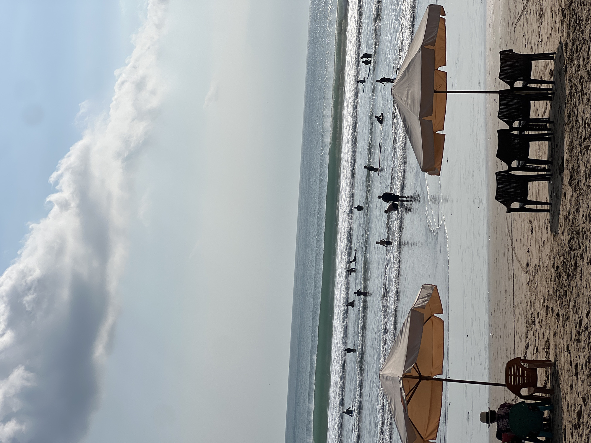 The image shows a beach scene in Kuta, Indonesia. Two beach umbrellas with attached chairs are set up on the sandy shore. In the foreground, one umbrella is partially visible, while the other is more fully in view, casting a shadow over several black plastic chairs. To the left, a person wearing a hat sits in a chair under the umbrella. In the middle ground, several people are in the ocean, some appearing to be surfing or learning to surf. The ocean has moderate waves, with a larger wave breaking further out. The sky above is a pale blue with scattered clouds. The overall lighting suggests it is daytime, possibly late afternoon.